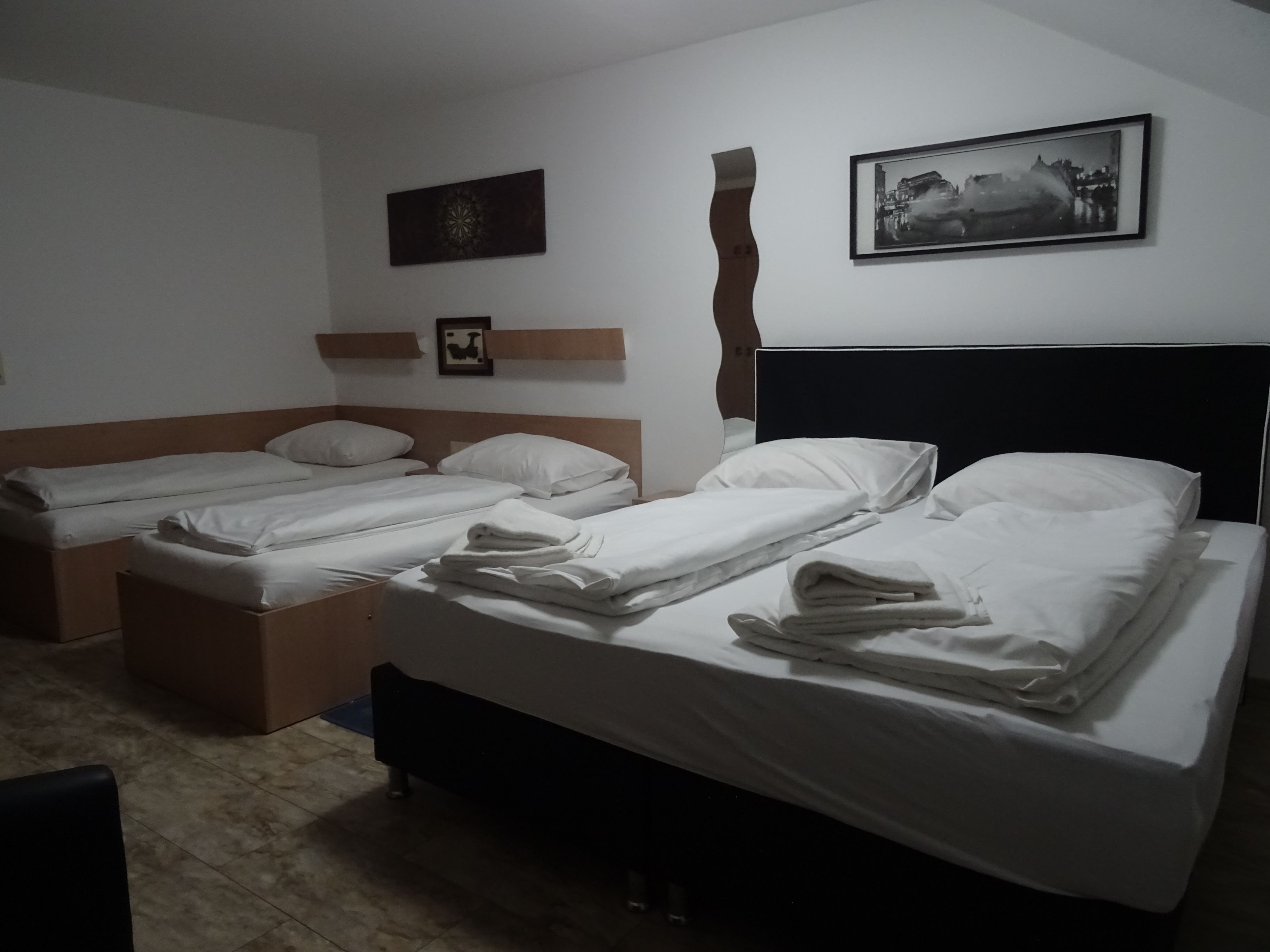 Une chambre d’hôtel avec deux lits simples et un lit double, tous équipés de draps blancs. On y trouve des cadres photos accrochés au mur et une étagère en bois sur laquelle est posée une image.
