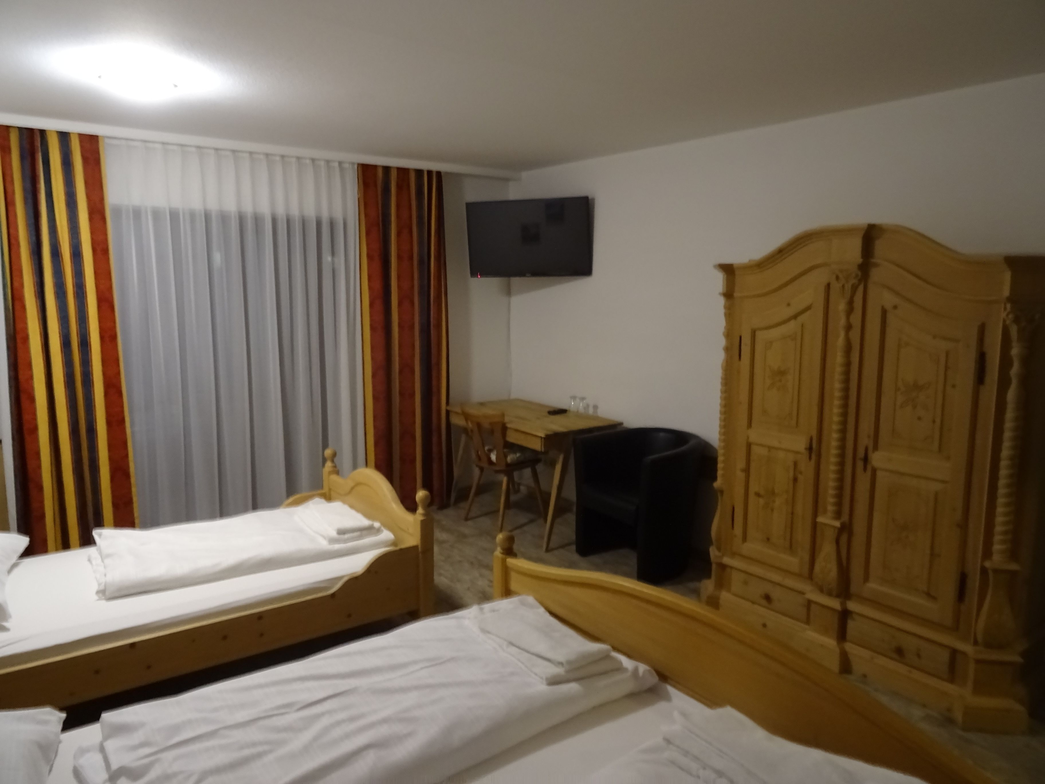 A hotel room with two single beds, a wooden wardrobe, a desk with a chair, and a television on the wall.