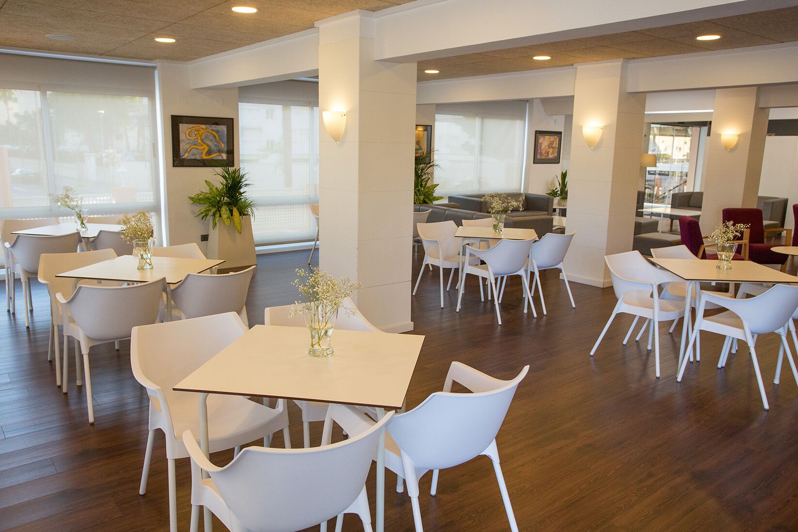 A modern dining area with white tables and chairs, large windows, and indoor plants.