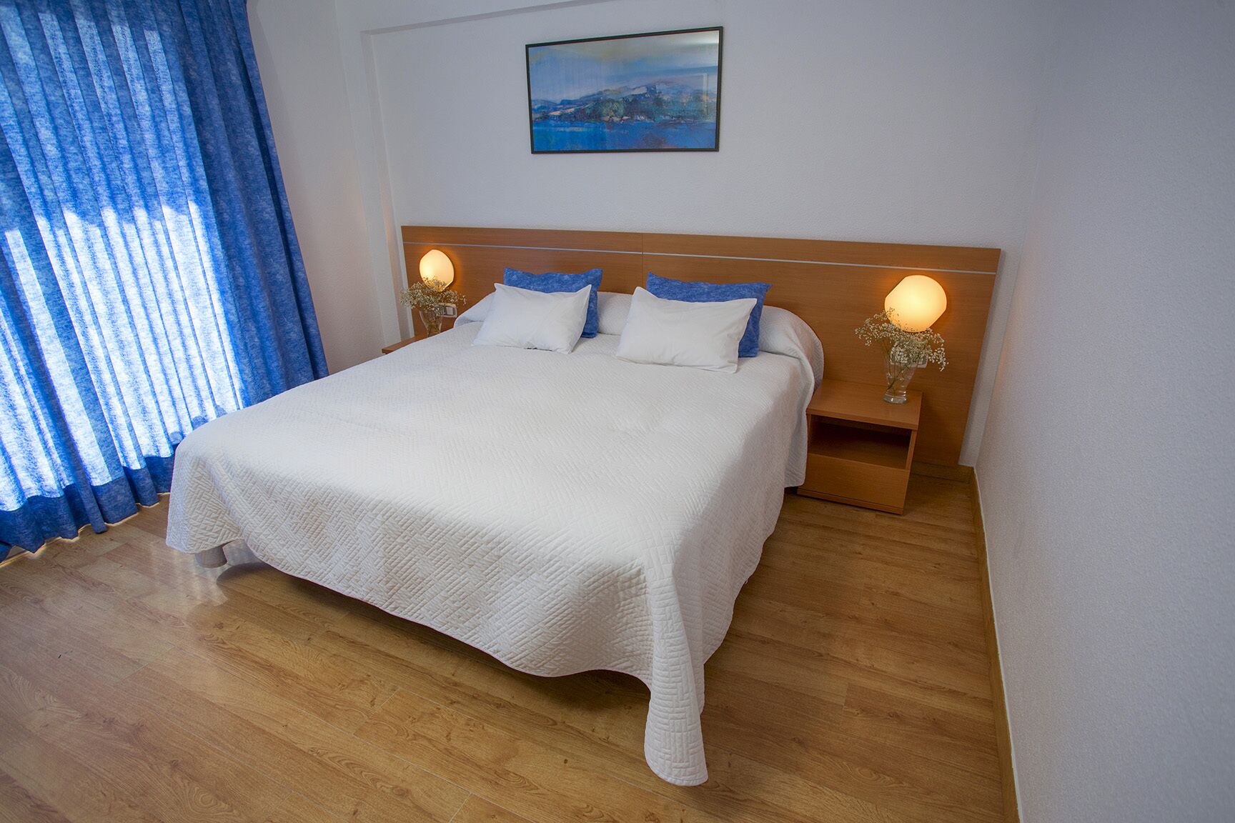 A neatly made bed with a white quilt, two blue pillows, and a wooden headboard. There are two bedside lamps with floral vases on the nightstands. A painting hangs above the bed.