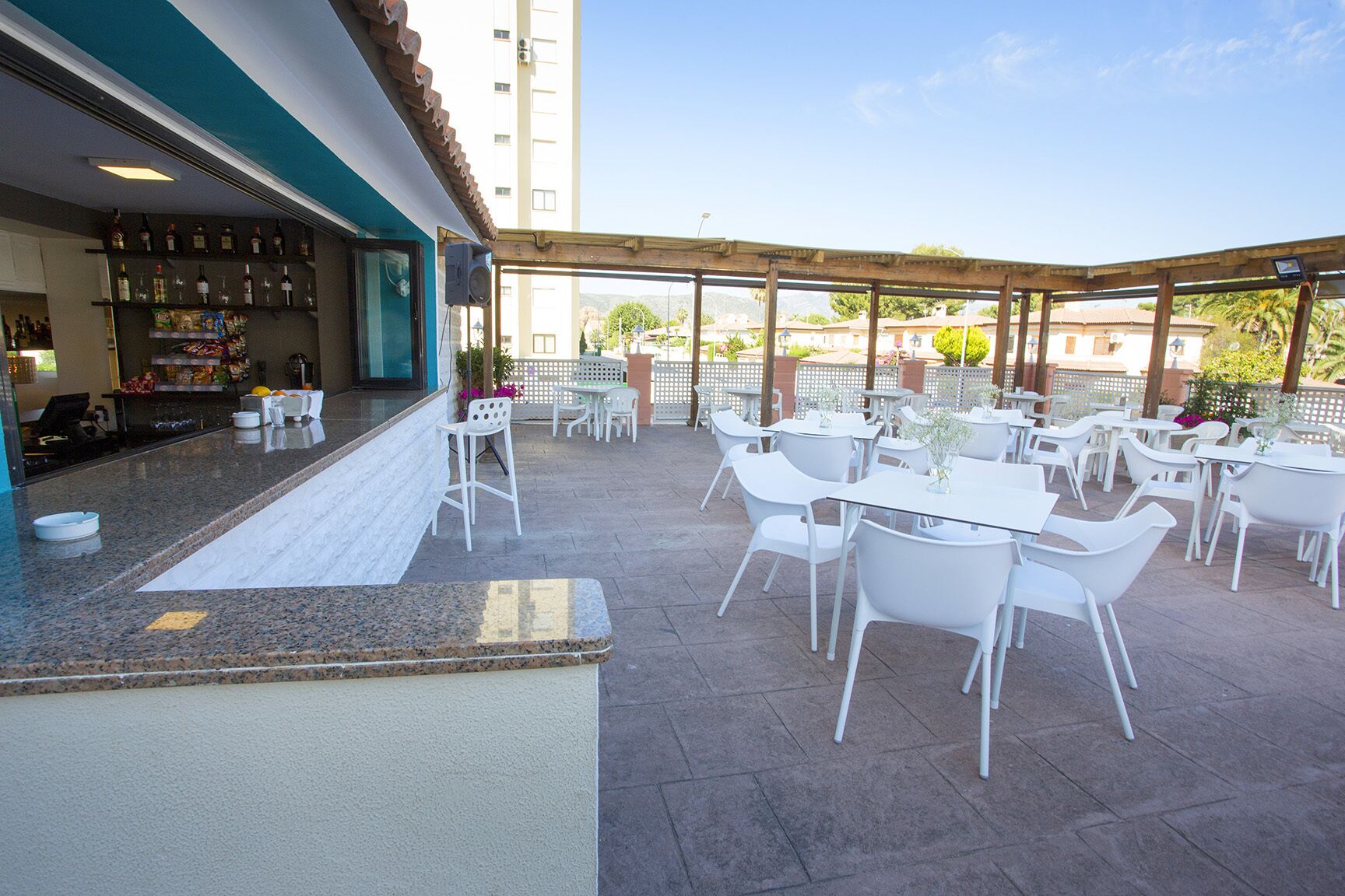 An outdoor patio area with white tables and chairs, a bar with shelves of bottles, and a view of buildings in the background.