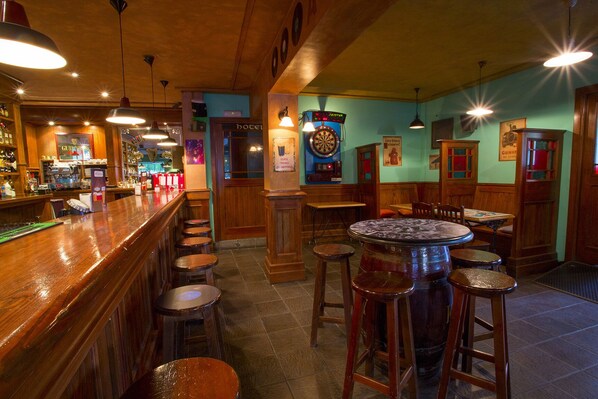 Zona de bar con mostradores de madera, taburetes y luces colgantes. Hay mesas y sillas para sentarse, además de un dardo en la pared.