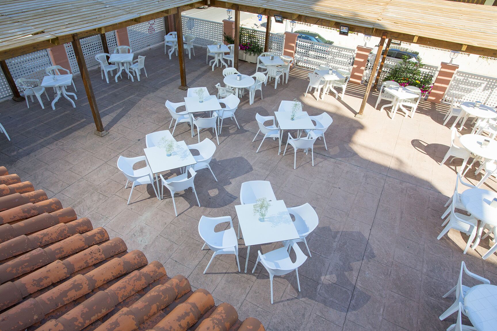 An outdoor patio area with multiple white tables and chairs, a covered section with a wooden roof, and a brick wall in the background.