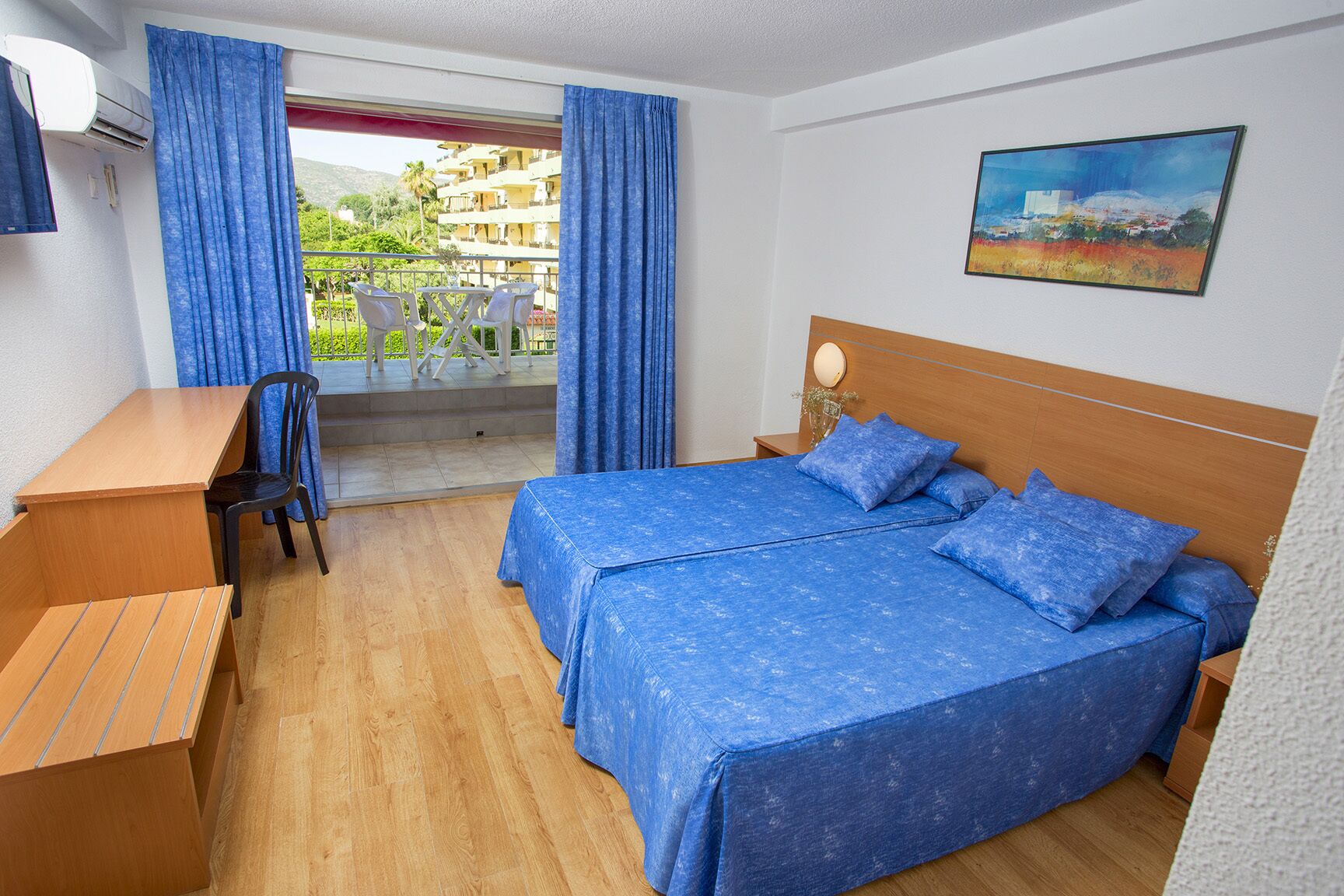 A hotel room with a wooden desk, two beds with blue bedding, a balcony with blue curtains, and a painting on the wall.