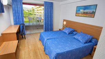 A hotel room with a wooden desk, two beds with blue bedding, a balcony with blue curtains, and a painting on the wall.