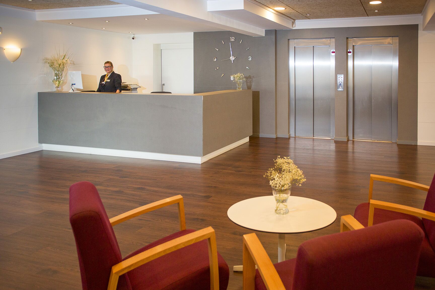 A hotel reception area with a receptionist, a reception desk, a clock, and two elevators.