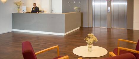 A hotel reception area with a receptionist, a reception desk, a clock, and two elevators.