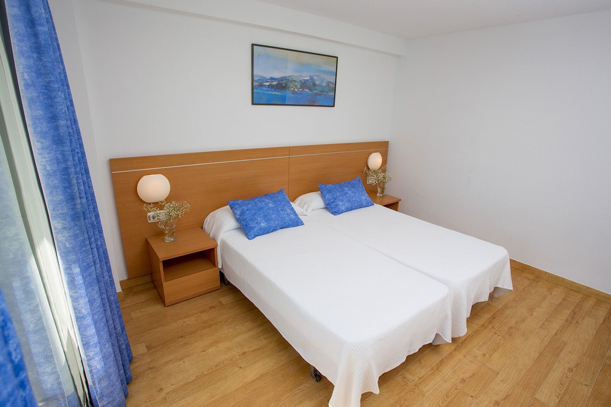 A neatly arranged bedroom with a wooden headboard, two bedside tables, a white bedspread, and a single blue pillow.