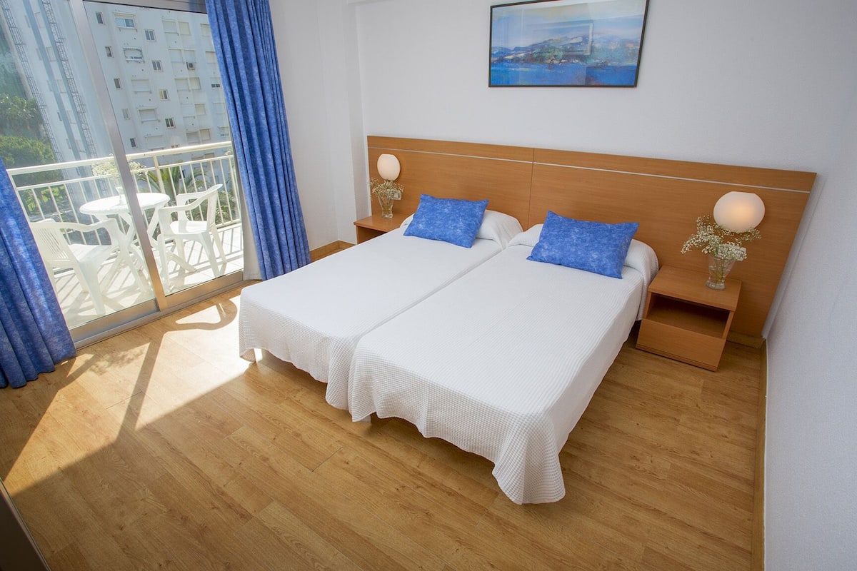 A double bed with white bedding and blue pillows, a wooden headboard, a bedside table with a lamp, and a window with blue curtains.