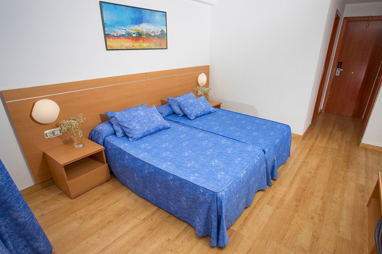 A double bed with a blue bedspread, wooden headboard, bedside tables with lamps, and a painting on the wall.