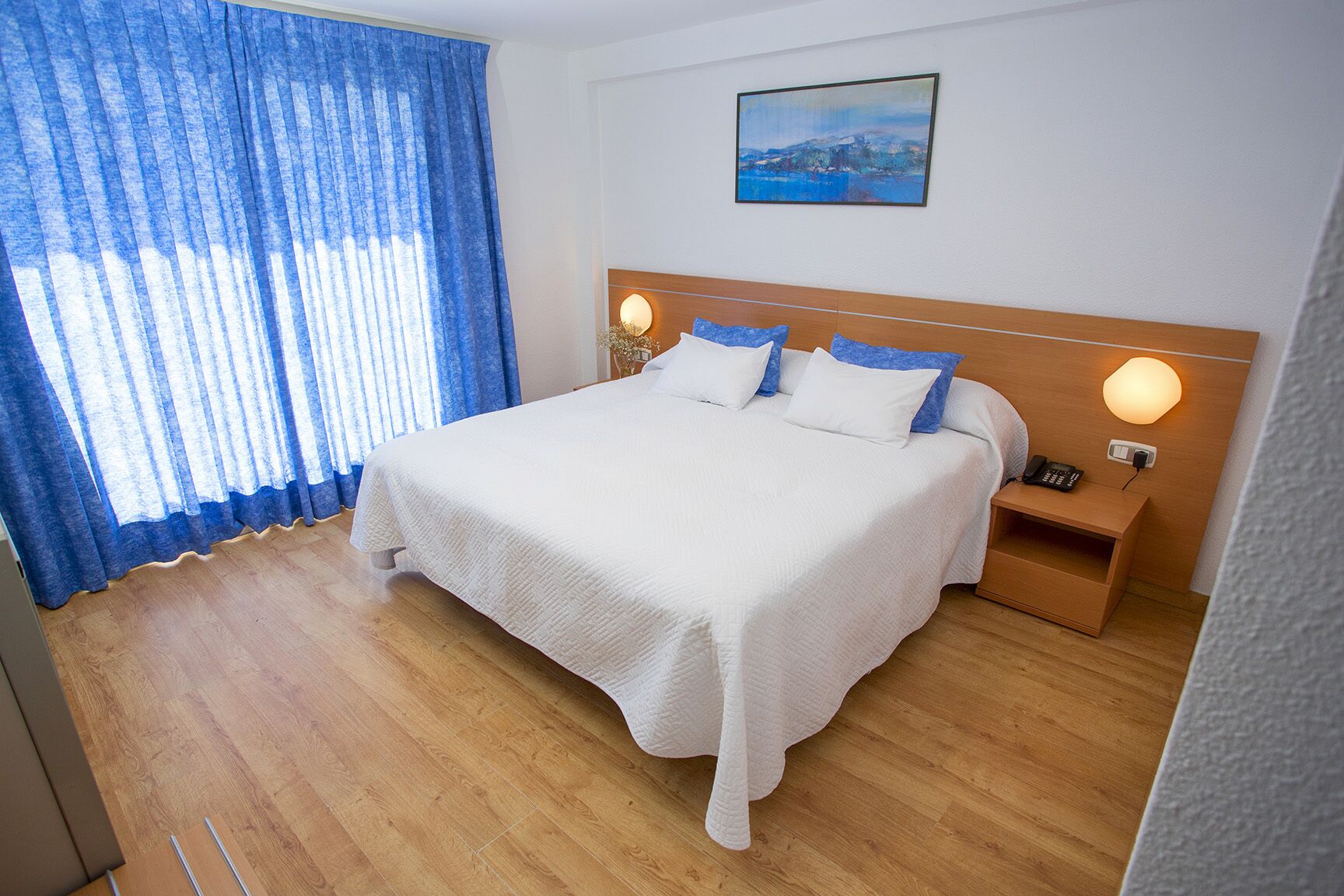 Una habitación de hotel con una cama grande, cabecera de madera, dos mesitas de noche con lámparas, un cuadro en la pared y una ventana con cortinas azules.
