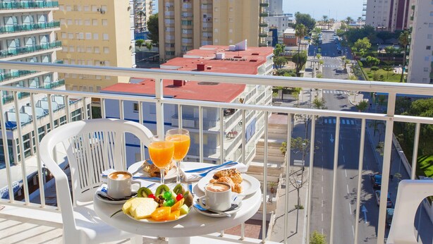Un balcón con una mesa dispuesta para el desayuno, incluyendo fruta, pasteles y café, con vista a un paisaje urbano con edificios altos y un cielo despejado.