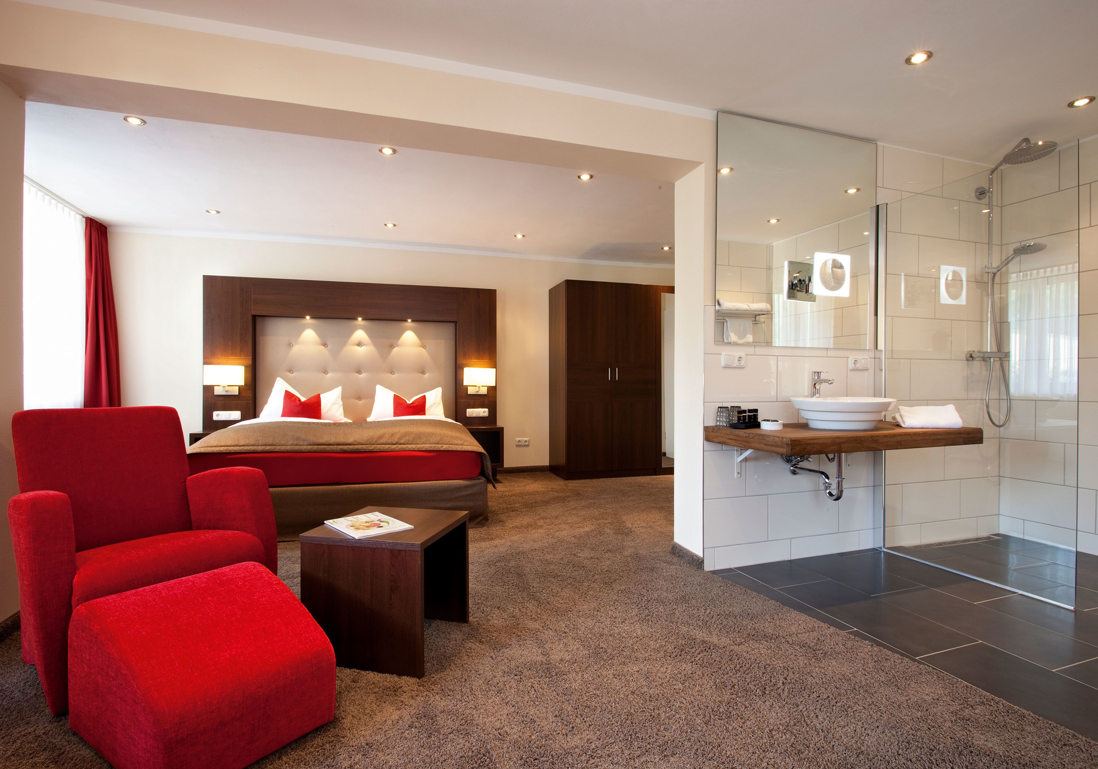 Ett modernt hotellrum med en stor säng, en röd fåtölj, ett litet bord, ett badrum med handfat och dusch samt en stor spegel.