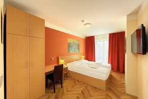 Quarto de hotel com cama, guarda-roupa de madeira, escrivaninha, cadeira e televisão.