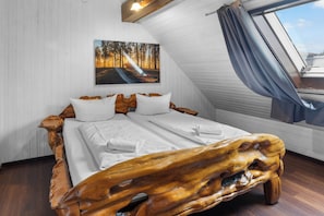 A bedroom with a wooden headboard, a bed with white linens, a painting of a forest on the wall, and a window with a view of the sky.