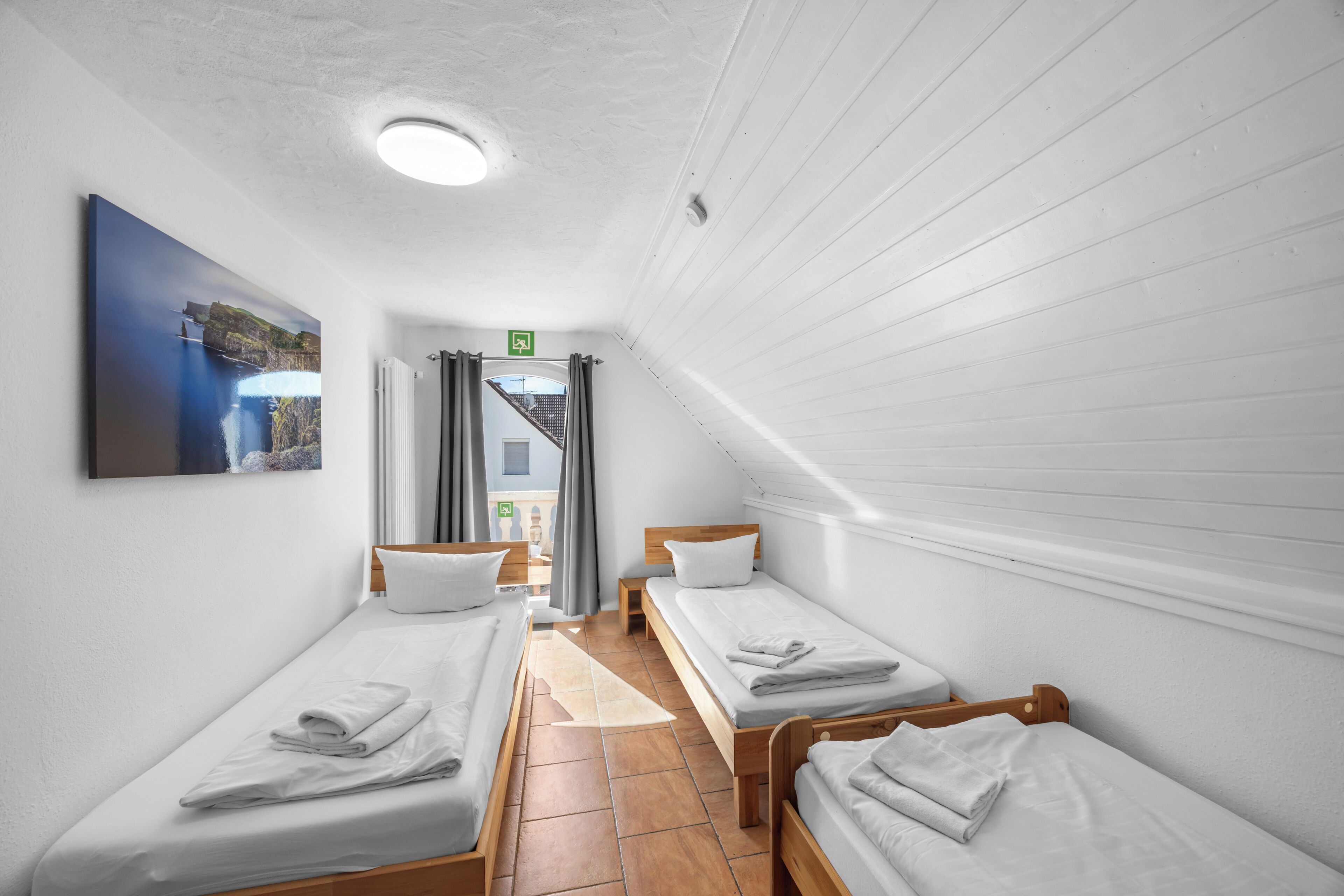 A room with three single beds, a wooden headboard, a white ceiling, and a view of a building through a window.