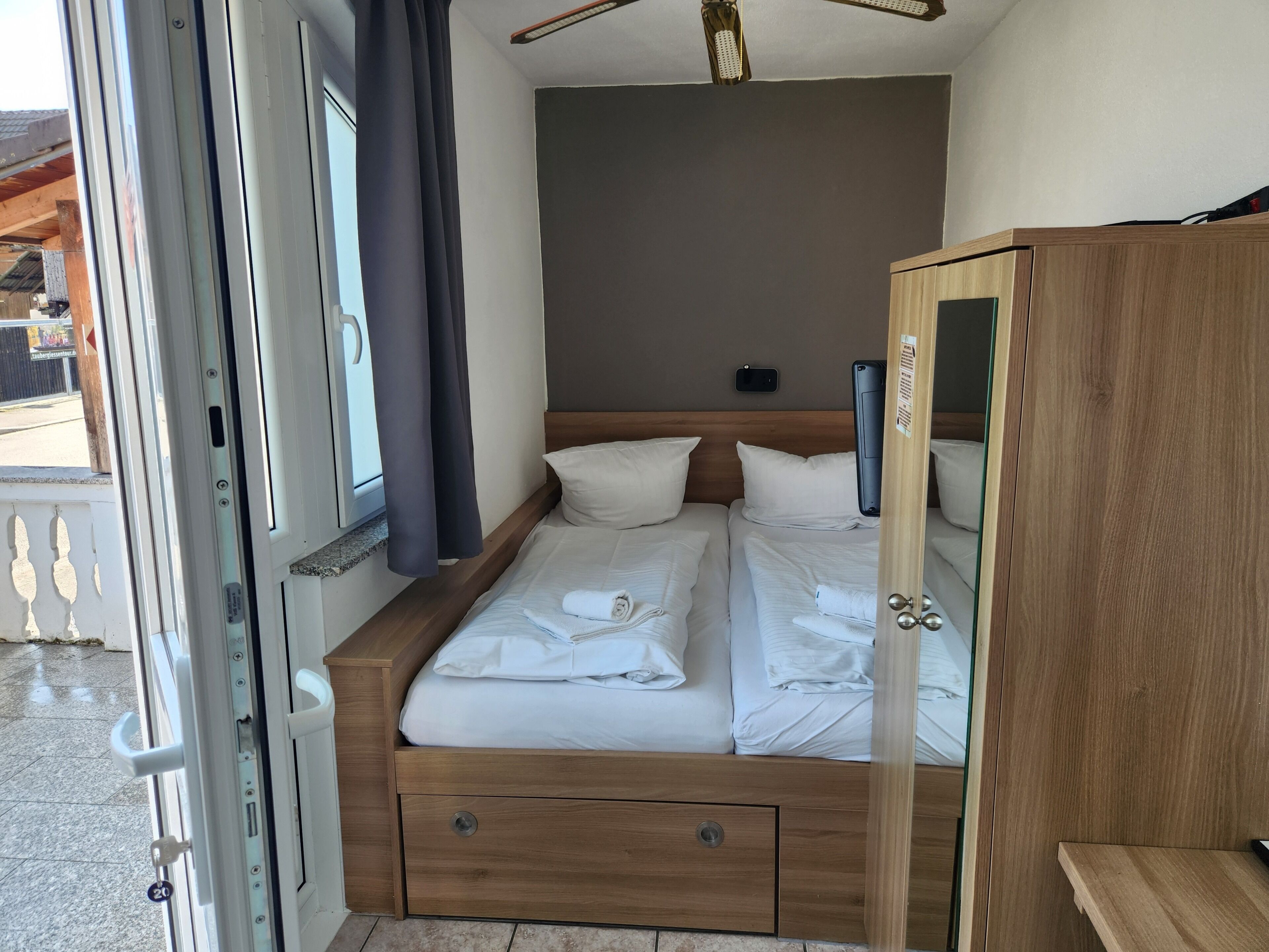 A hotel room with a bed, a wooden wardrobe, a bedside table, and a window with curtains.
