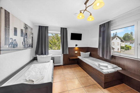 Une chambre avec deux lits, un téléviseur, une fenêtre avec des rideaux et un luminaire fixé au mur.