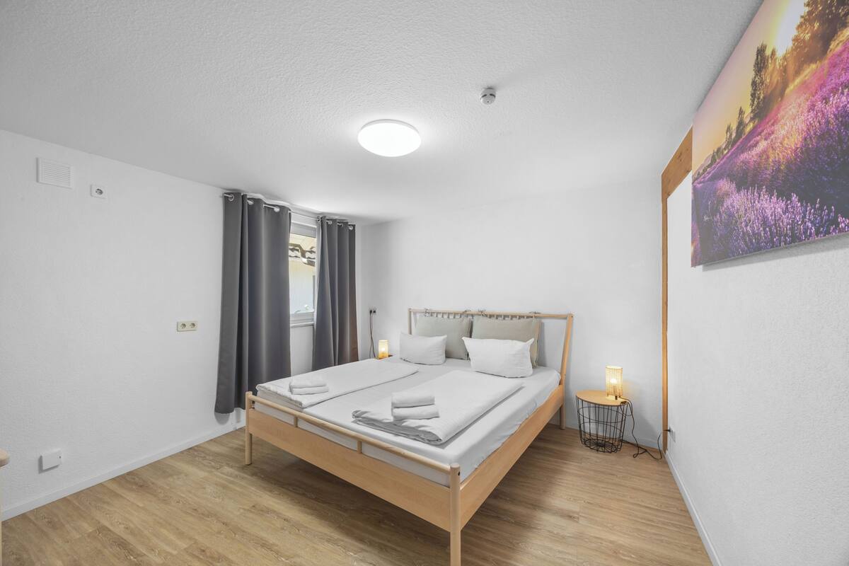 A bedroom with a wooden bed, a bedside table with a lamp, a window with curtains, and a wall-mounted picture.