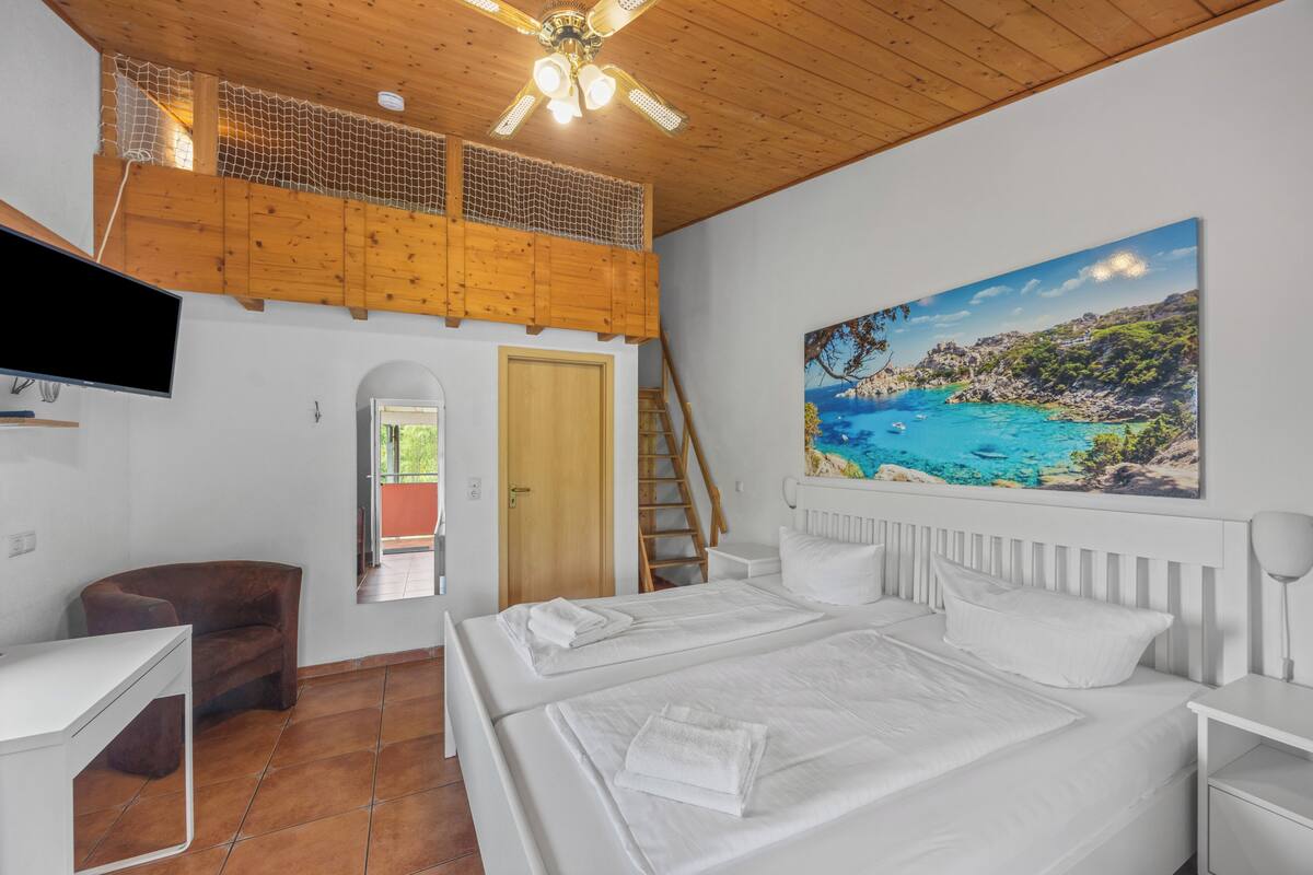 A bedroom with a bed, a desk, a chair, a painting, and a balcony with a wooden railing.