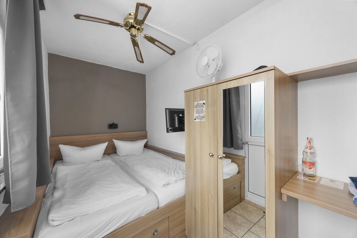A bedroom with a bed, wooden wardrobe, ceiling fan, and a small table with a bottle.