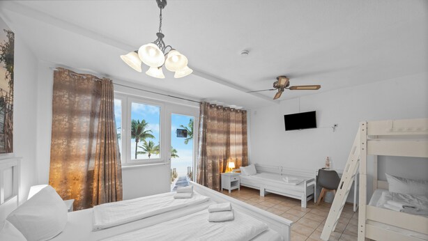 Habitación de hotel con cama, escritorio, silla, televisor, literas y vistas a palmeras.