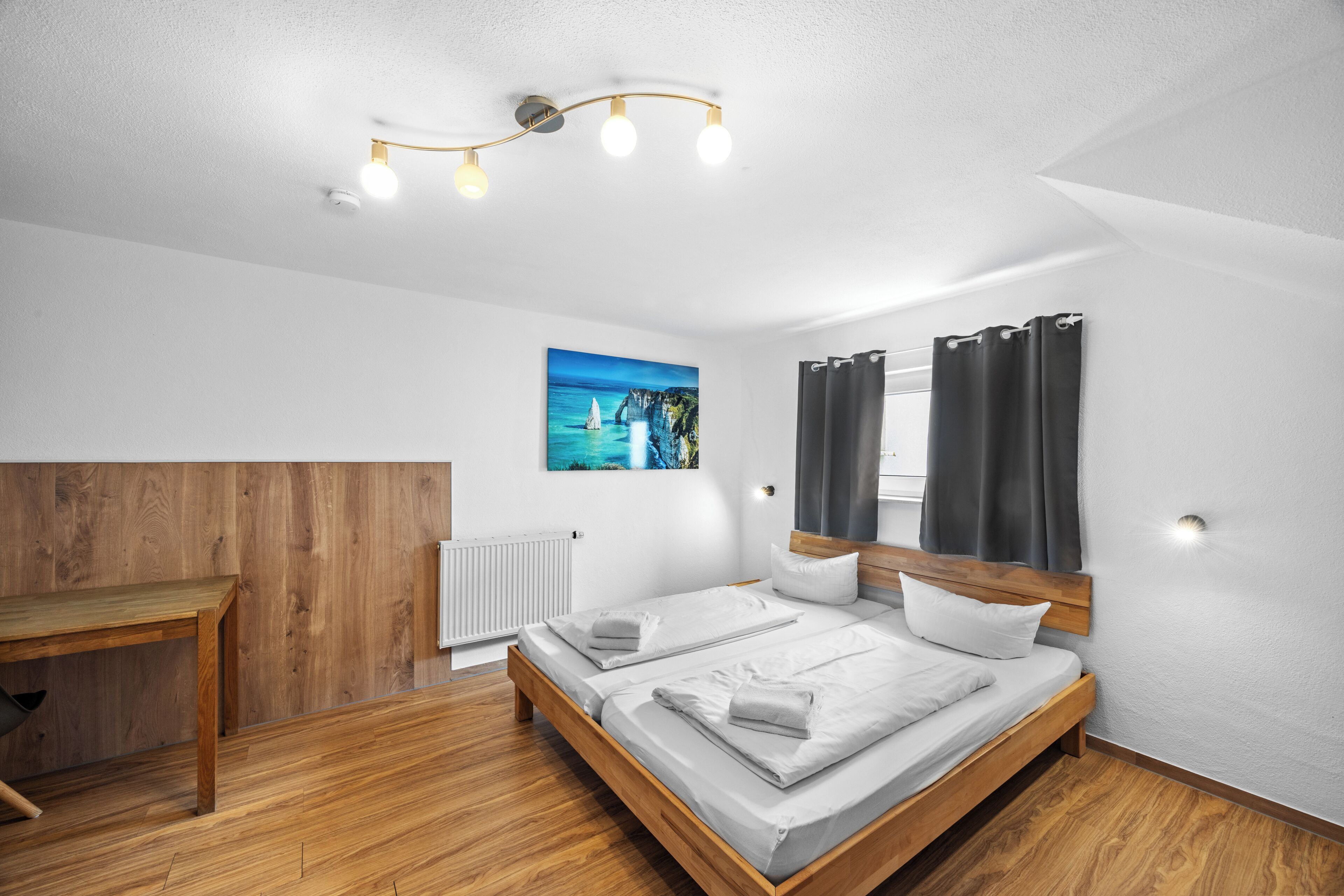 Ein modernes Schlafzimmer mit einem hölzernen Bett, einem kleinen Tisch, einem Gemälde einer Küstenlandschaft und einer Deckenleuchte.