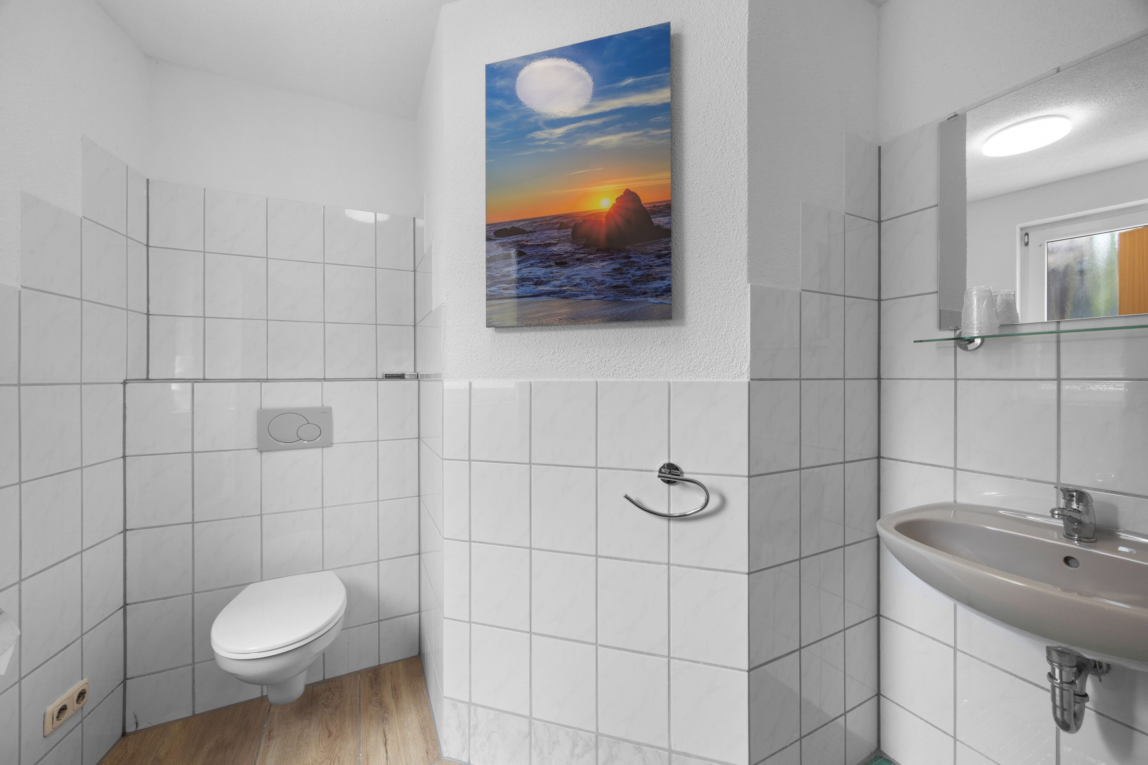 Une salle de bain avec des carreaux blancs, des toilettes, un lavabo et un tableau représentant un coucher de soleil sur l’océan.