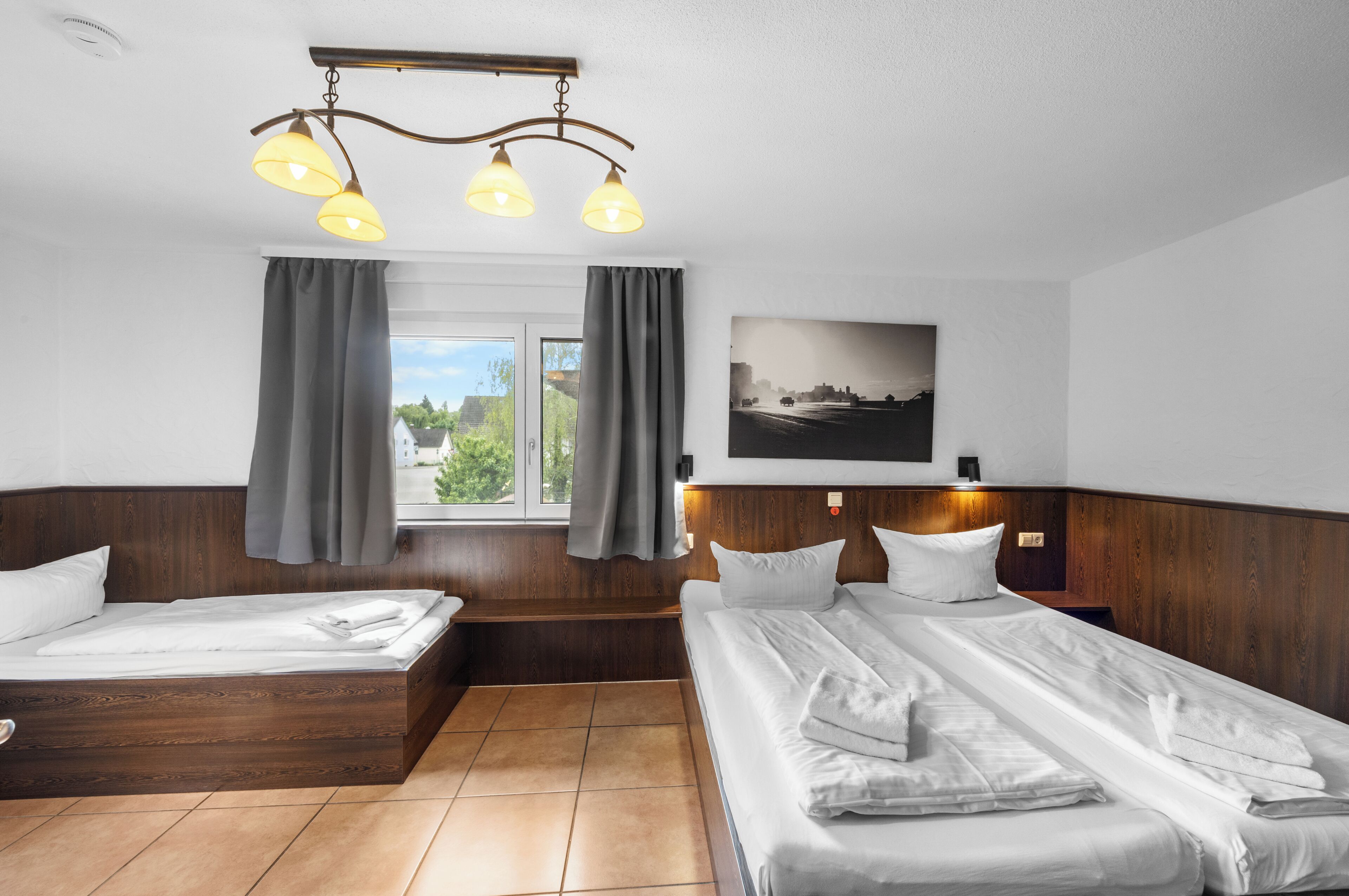 Ein Hotelzimmer mit zwei Betten, einem großen Fenster mit Vorhängen, einem Stadtbildgemälde und einer Deckenleuchte.