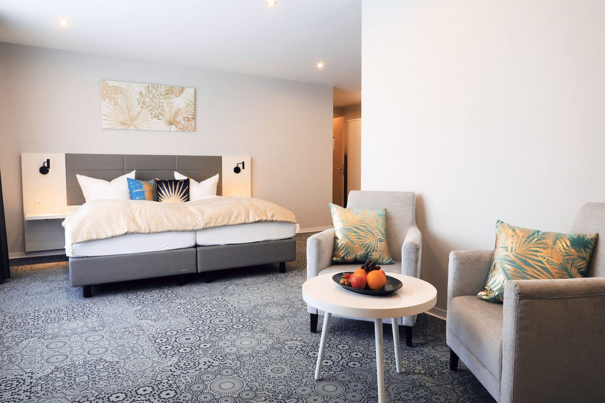 A modern hotel room with a grey sofa bed, two armchairs, a round coffee table with fruit, and a patterned carpet.