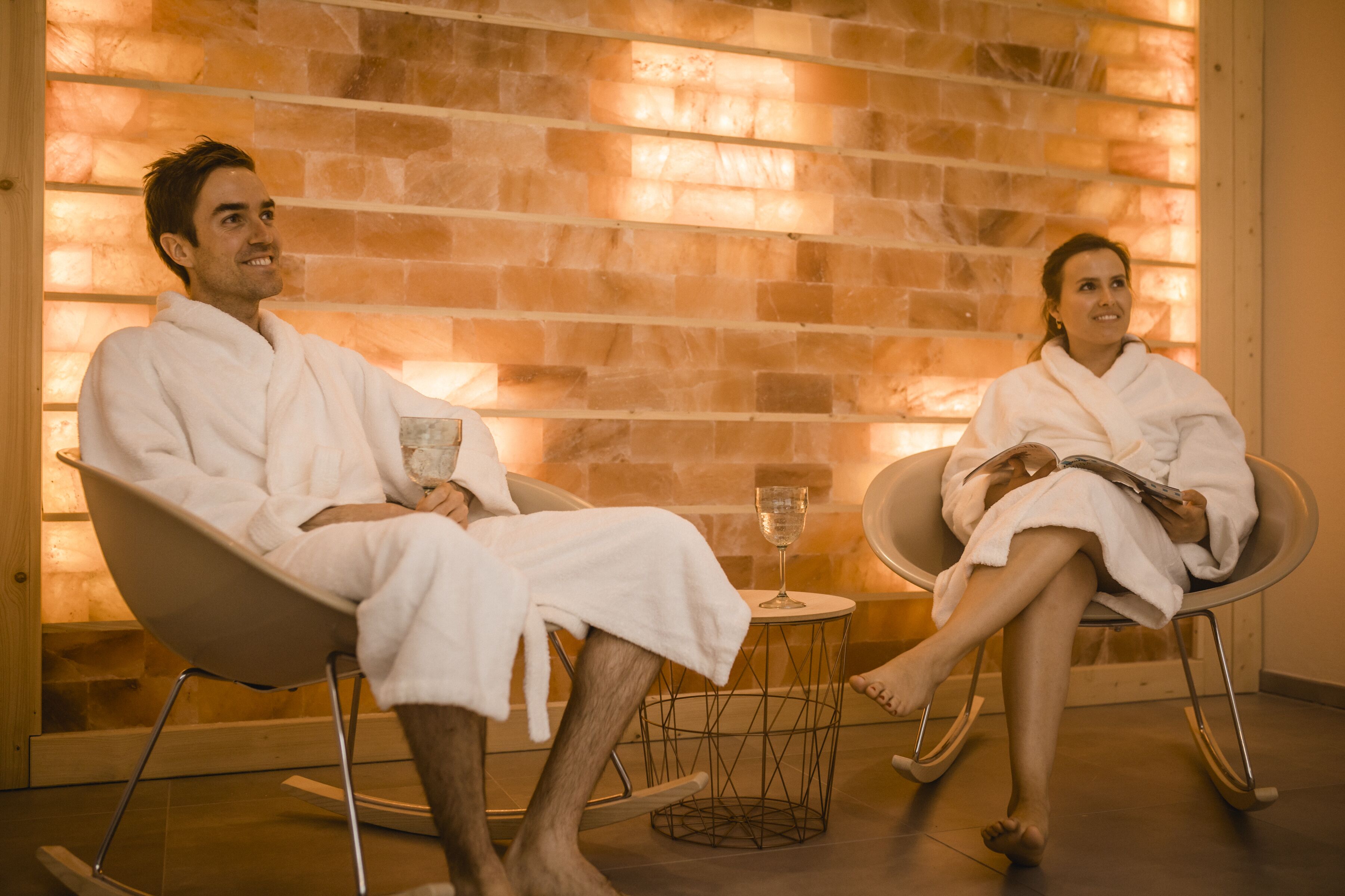 Two people in white robes seated in modern chairs in a room with a brick wall and a small table with a glass.
