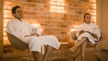 Two people in white robes seated in modern chairs in a room with a brick wall and a small table with a glass.
