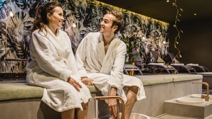 Una pareja con batas blancas sentada en un banco en un moderno balneario con un amplio mural de fondo y un diseño minimalista.