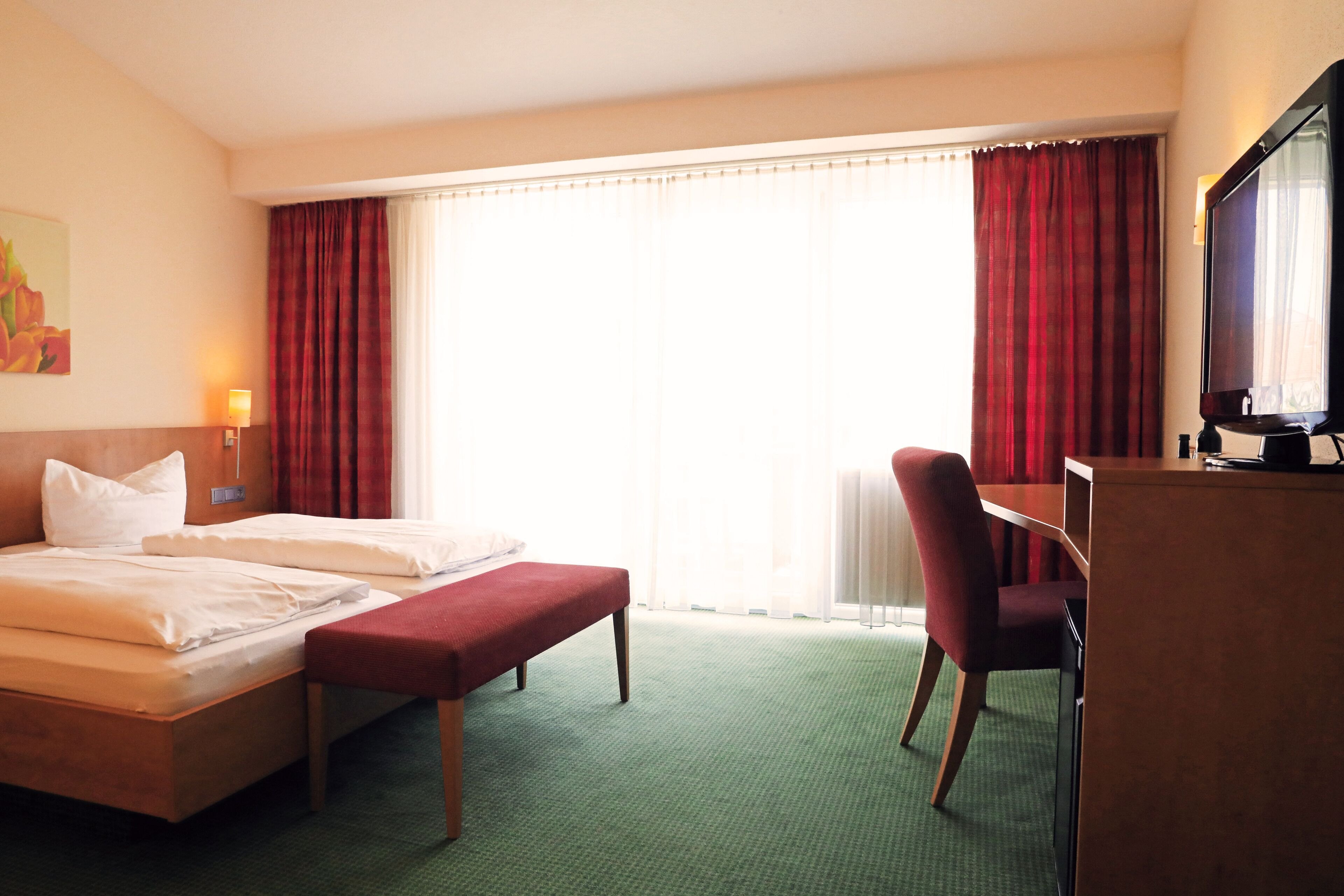 Ein Hotelzimmer mit Bett, Schreibtisch, Sessel, Fernseher und einem Fenster mit Vorhängen.
