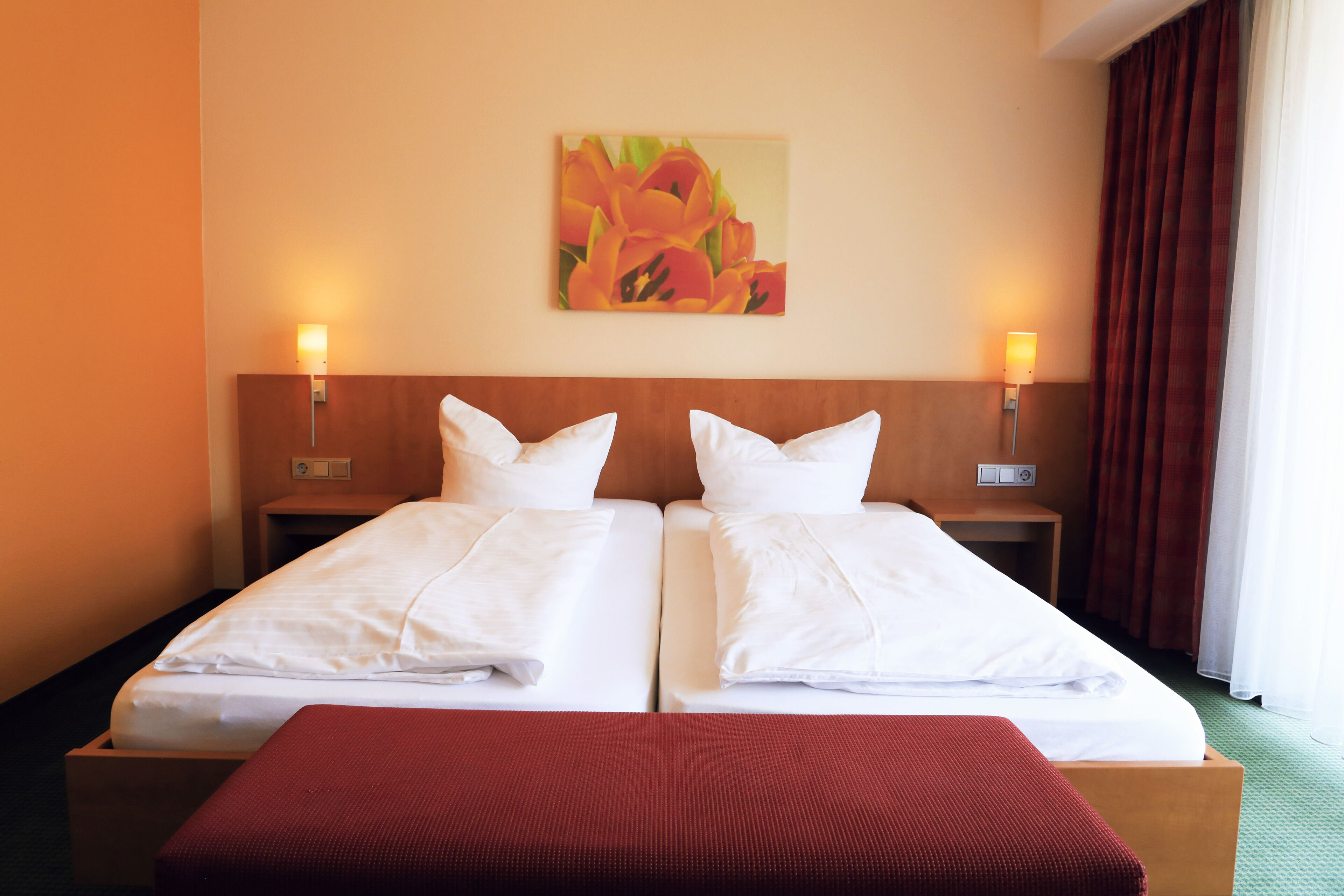 Ein Doppelbett mit weißen Bettwäsche und roter Decke, zwei Nachttische mit Lampen, ein Blumenbild an der Wand und ein Fenster mit Vorhängen.