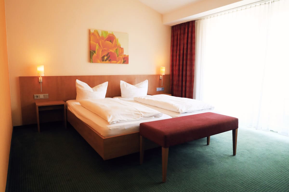 A hotel room with a double bed, two bedside tables, a bench, a painting on the wall, and a window with curtains.