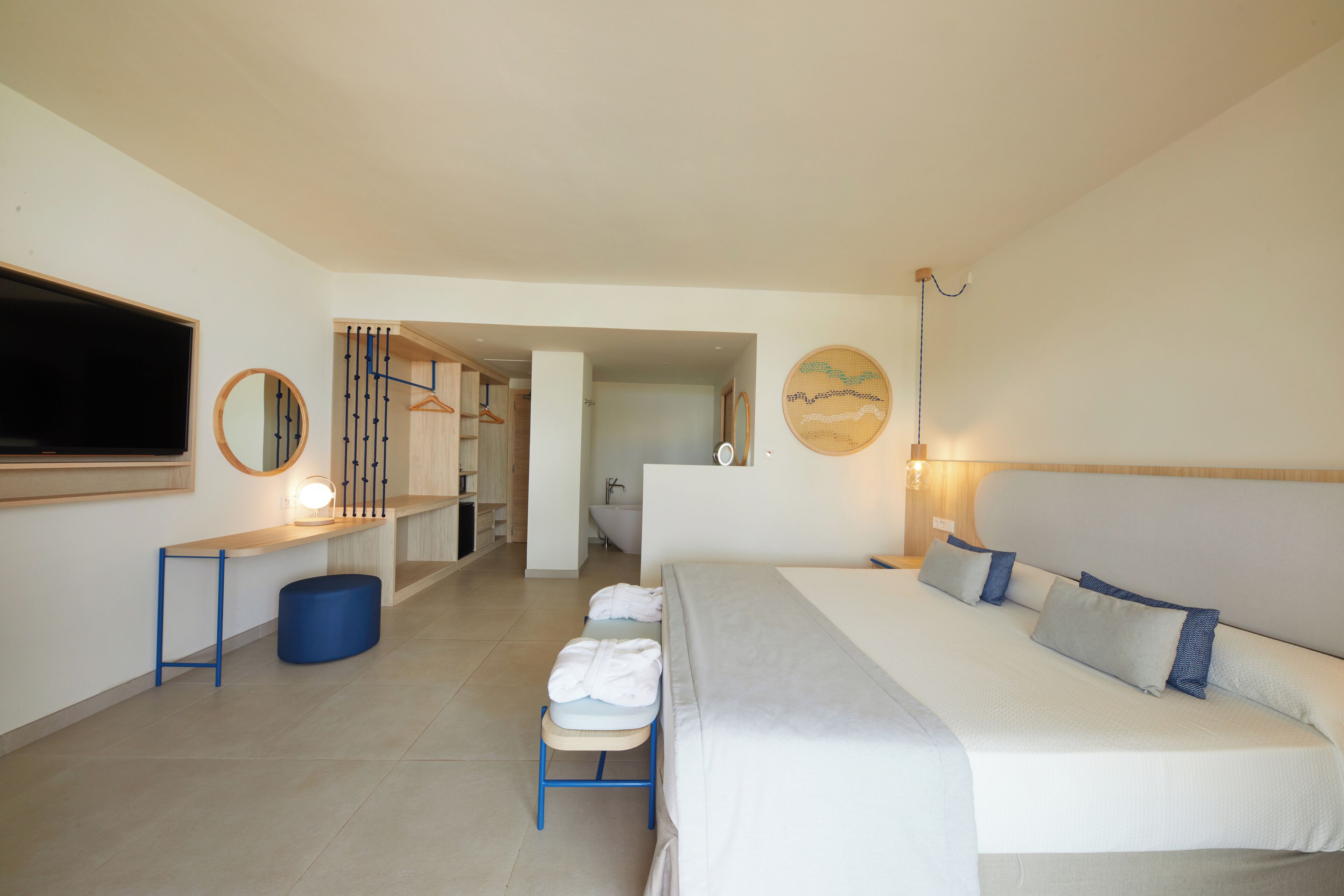 A hotel room with a large bed, a TV, a mirror, a small stool with towels, and a bathroom visible through an open door.