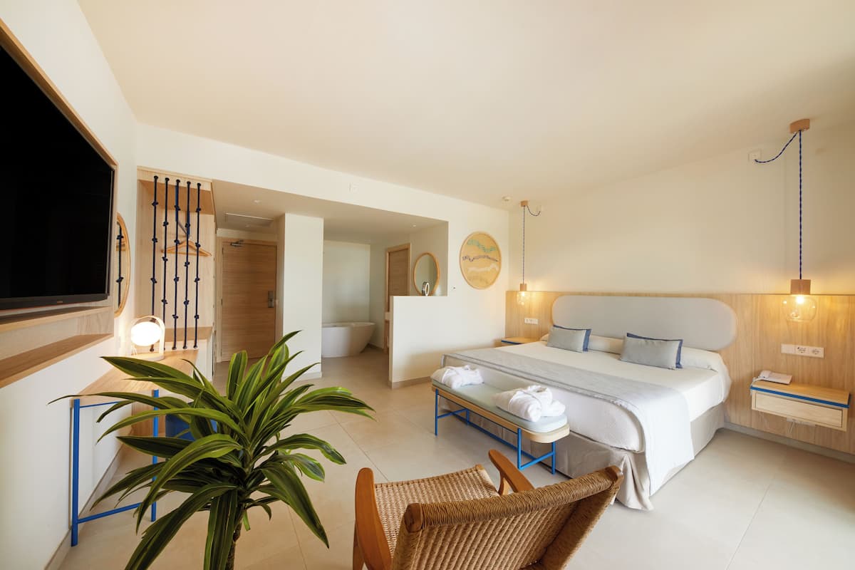 Habitación de hotel moderna con cama, mesita de noche, lámpara, planta en maceta y vista al pasillo.