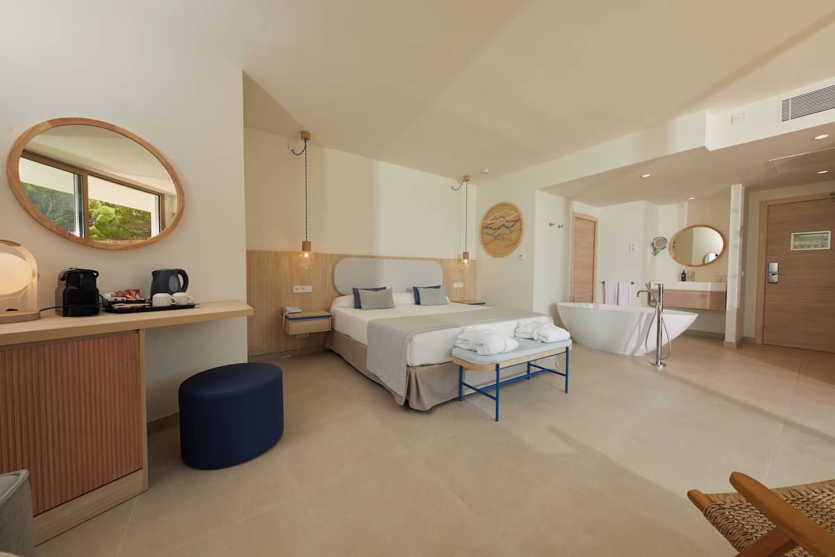Una habitación de hotel moderna con una cama grande, un escritorio con cafetera, un espejo redondo y una bañera.