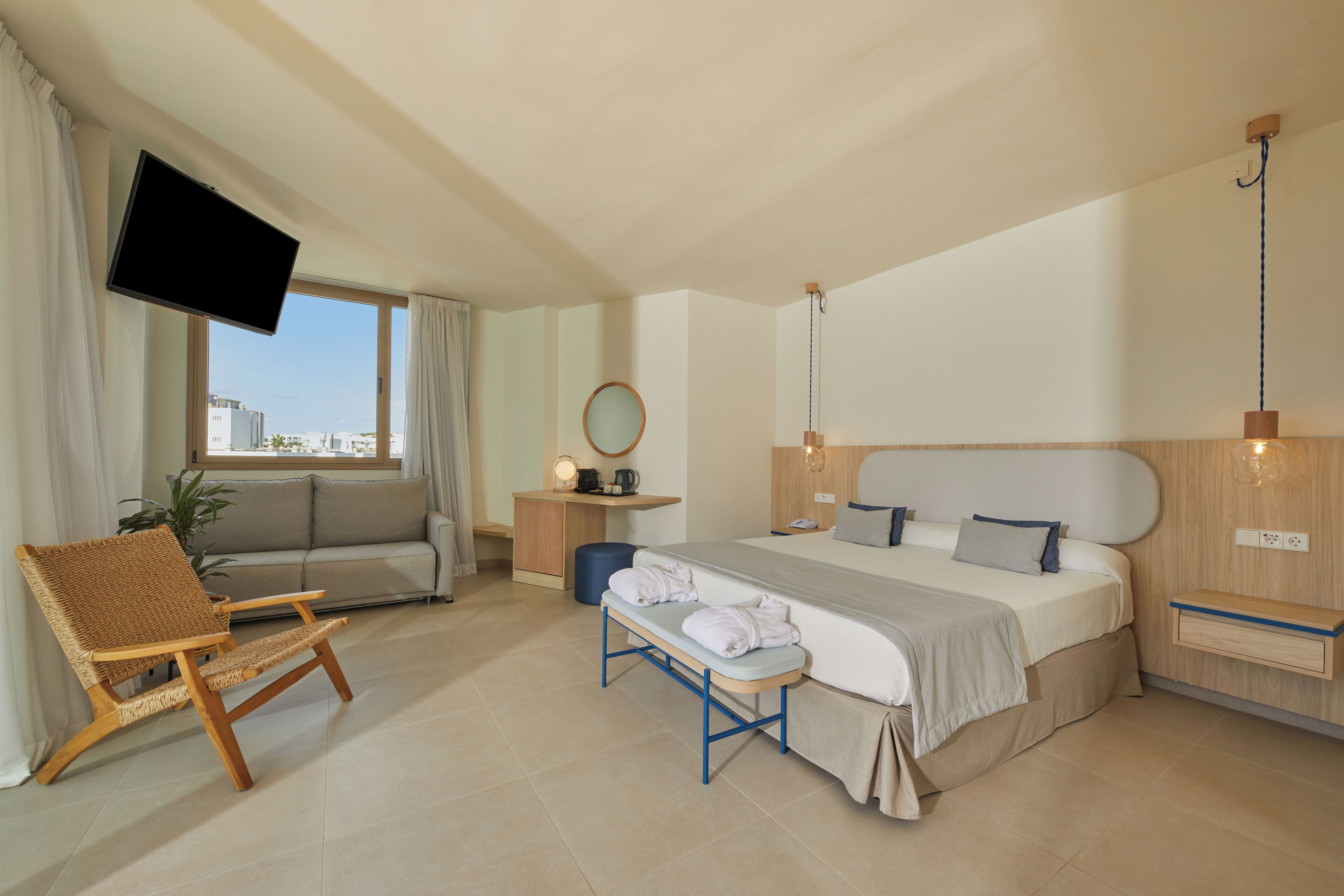 Una habitación de hotel moderna con una cama grande, una televisión montada en la pared, un área de descanso con una silla y un banco, y vistas de la ciudad a través de la ventana.