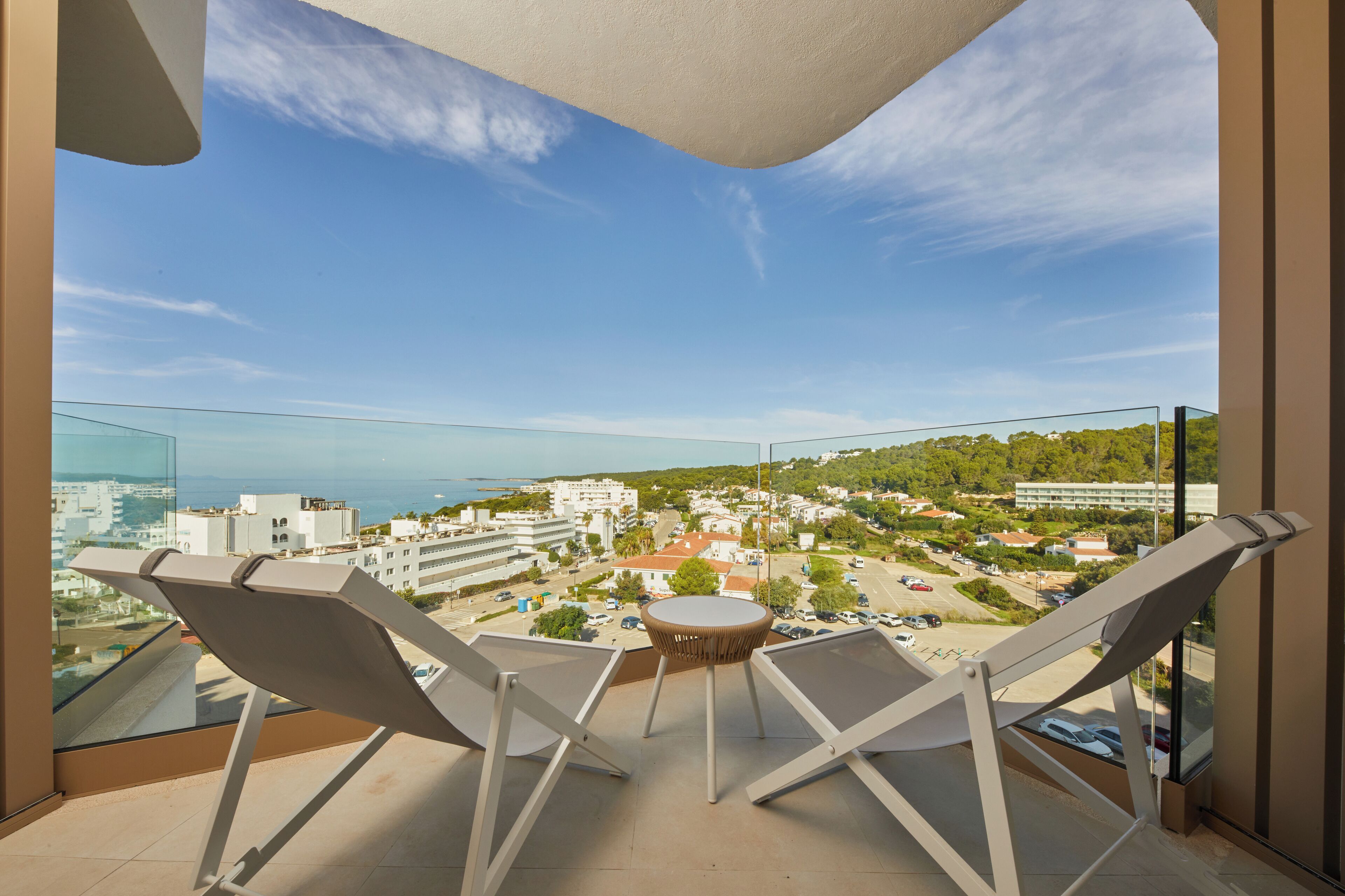 A balcony with two lounge chairs and a small table, overlooking a coastal town with buildings and greenery.