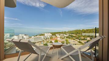 A balcony with two lounge chairs and a small table, overlooking a coastal town with buildings and greenery.