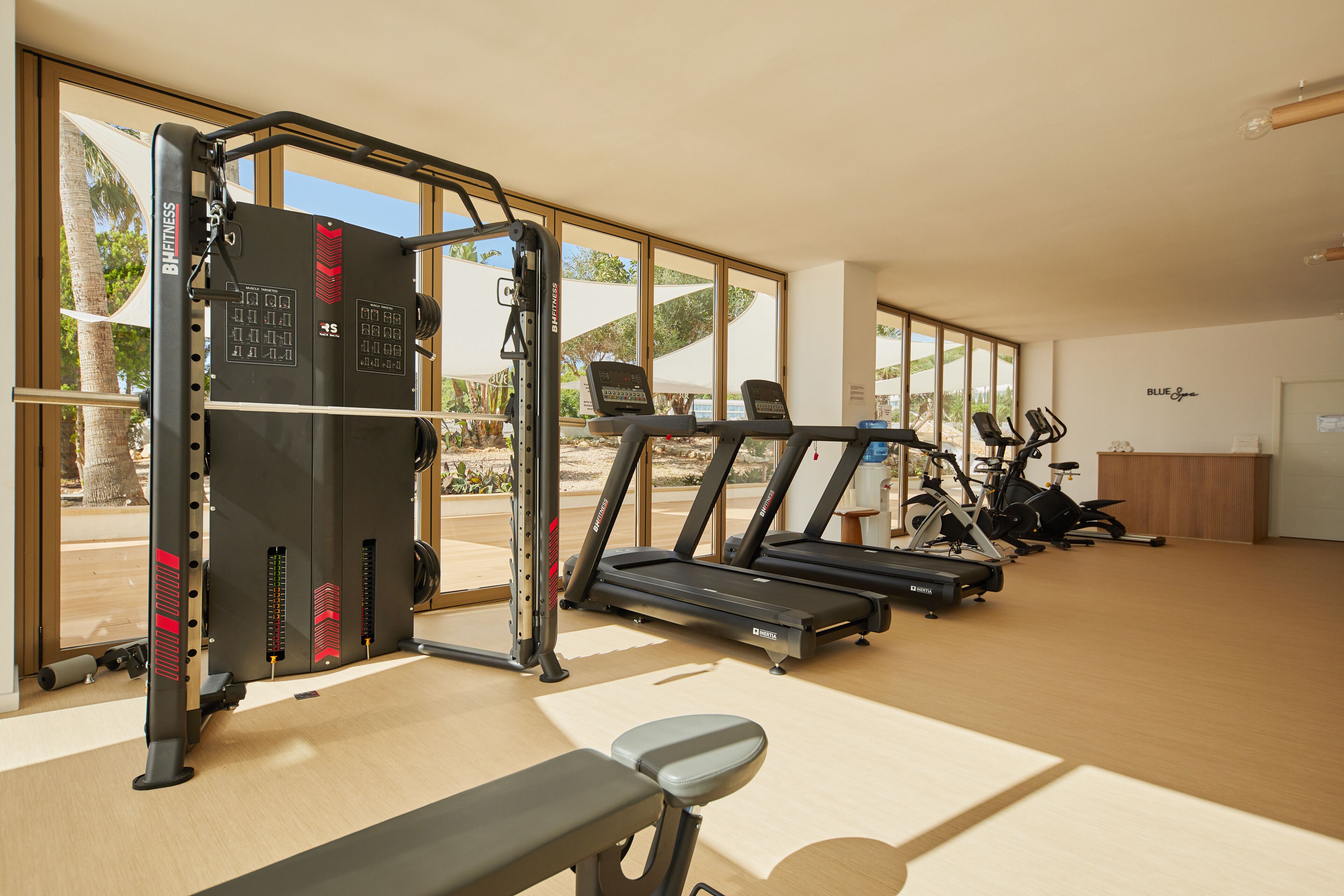 Un gimnasio con equipo de ejercicio, incluyendo cintas de correr y una máquina de pesas, con ventanas amplias que ofrecen vistas al exterior.