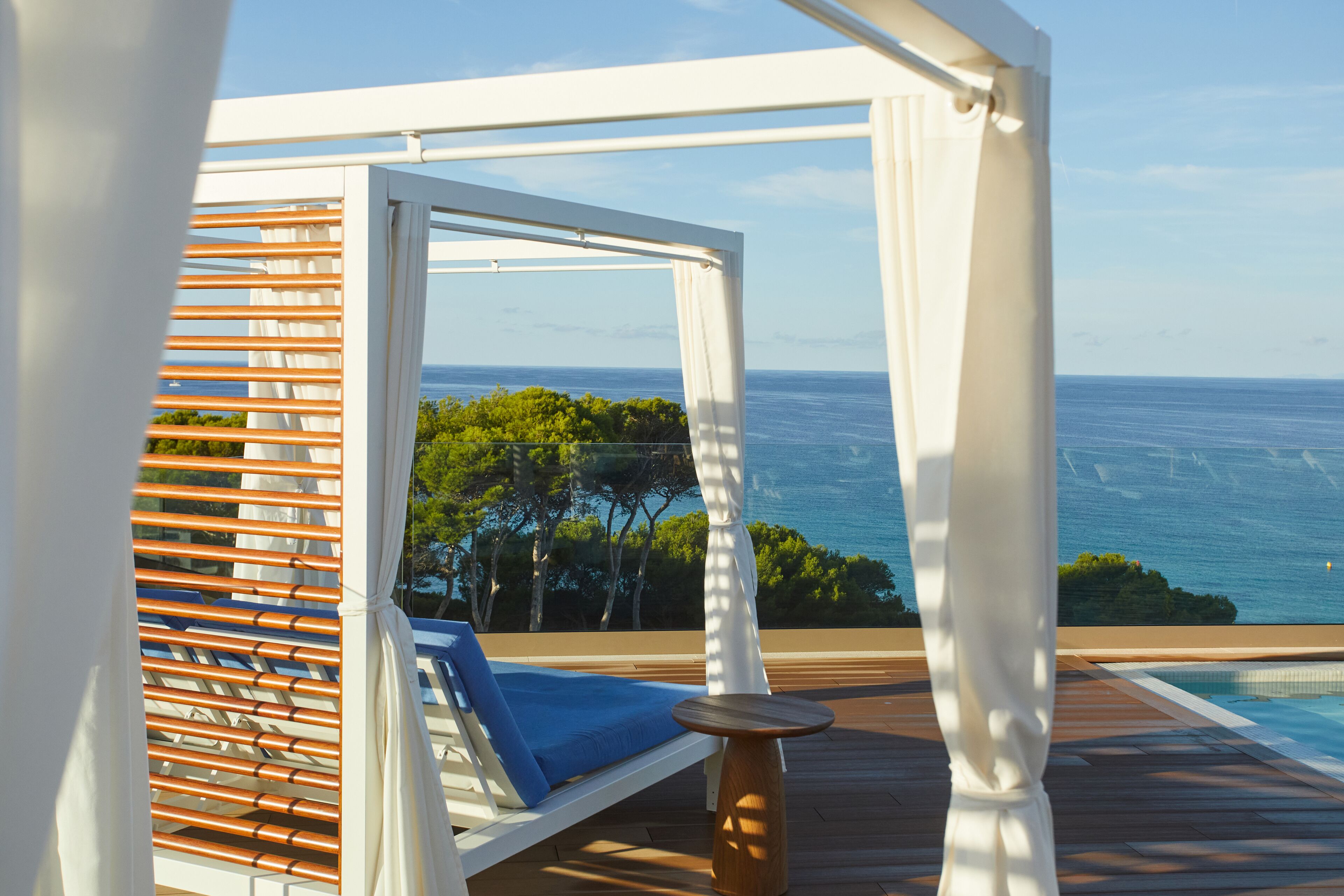 A sunlit outdoor lounge area with a pool, wooden deck, and a view of the ocean.