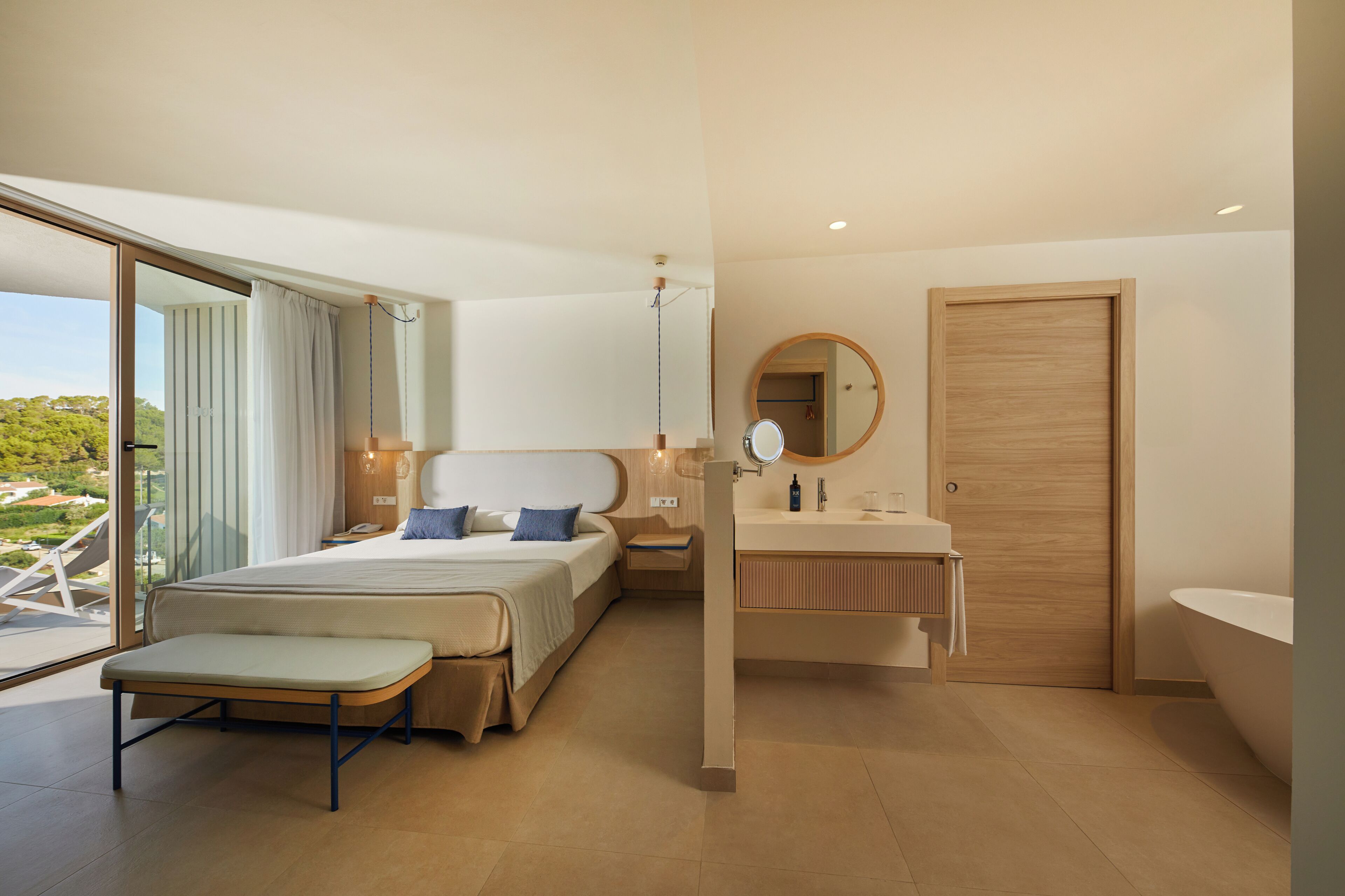 A modern hotel room with a large bed, a bedside table, a vanity with a mirror, and a bathtub.