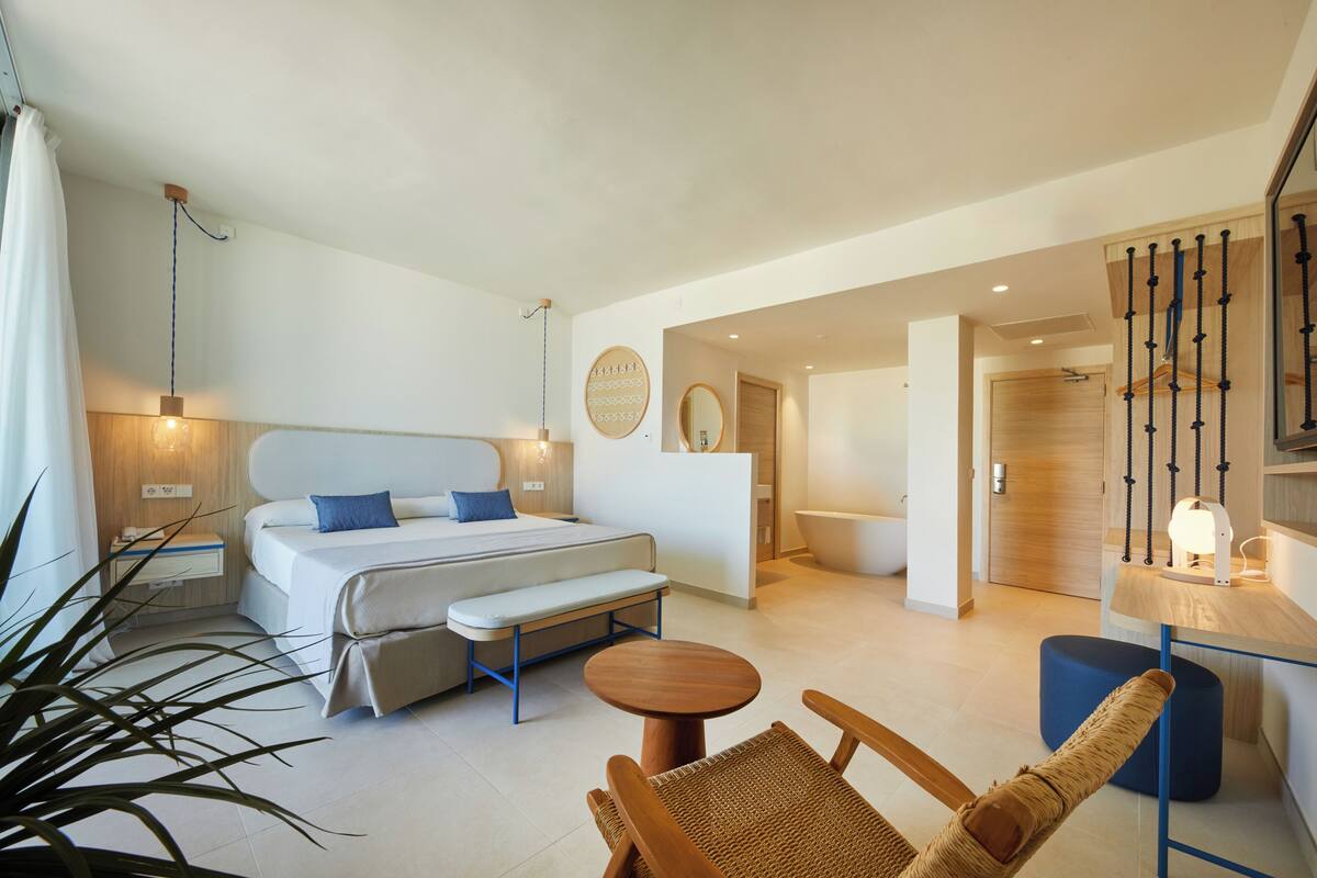 Una habitación de hotel moderna con una cama, mesita de noche, banco y una pequeña mesa redonda.
