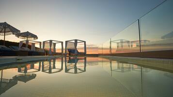 A clear pool with lounge chairs and umbrellas, surrounded by glass walls reflecting the sunset.