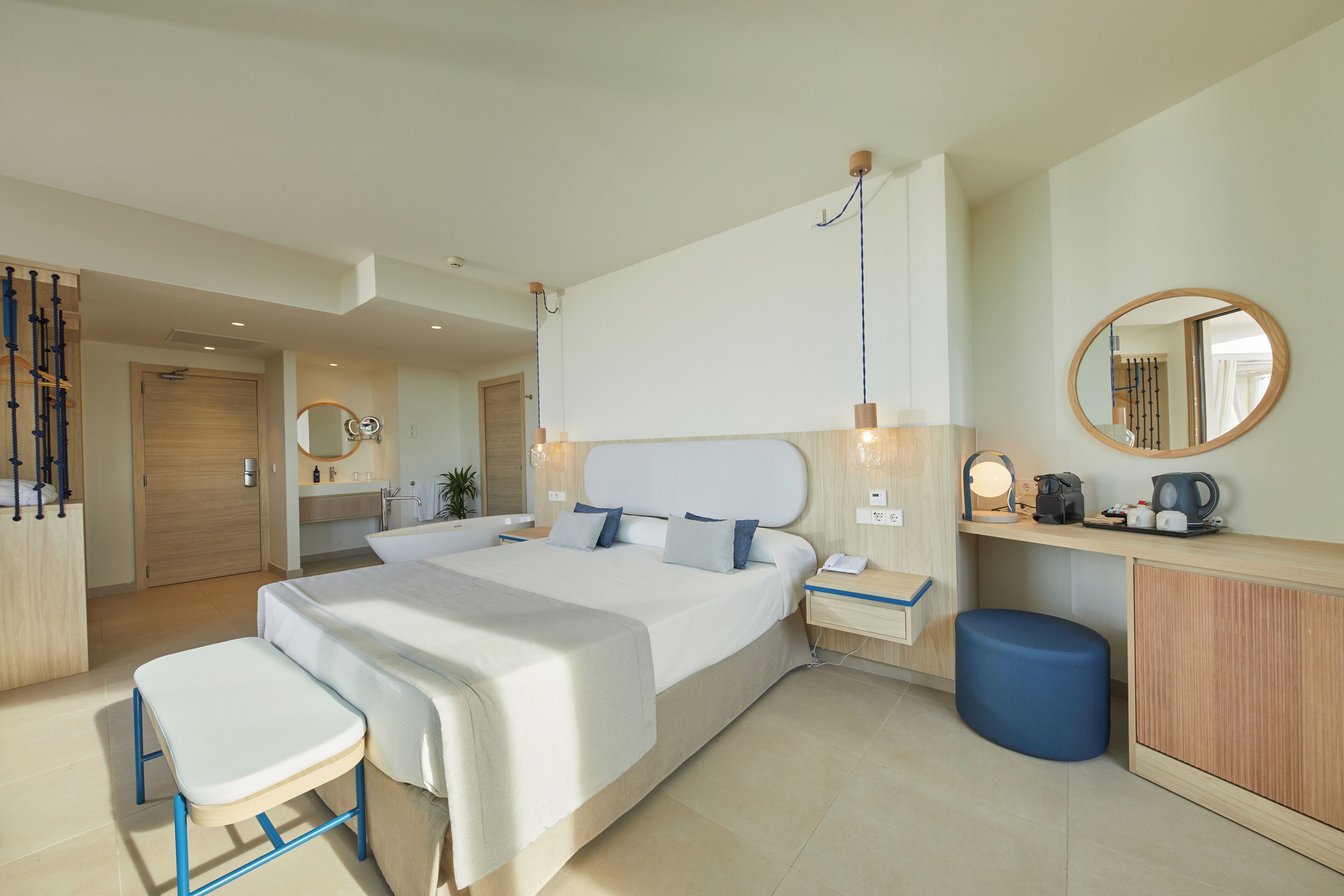 Una habitación de hotel moderna con una cama grande, mesitas de noche, un escritorio, una silla, un espejo y un reposapiés azul.