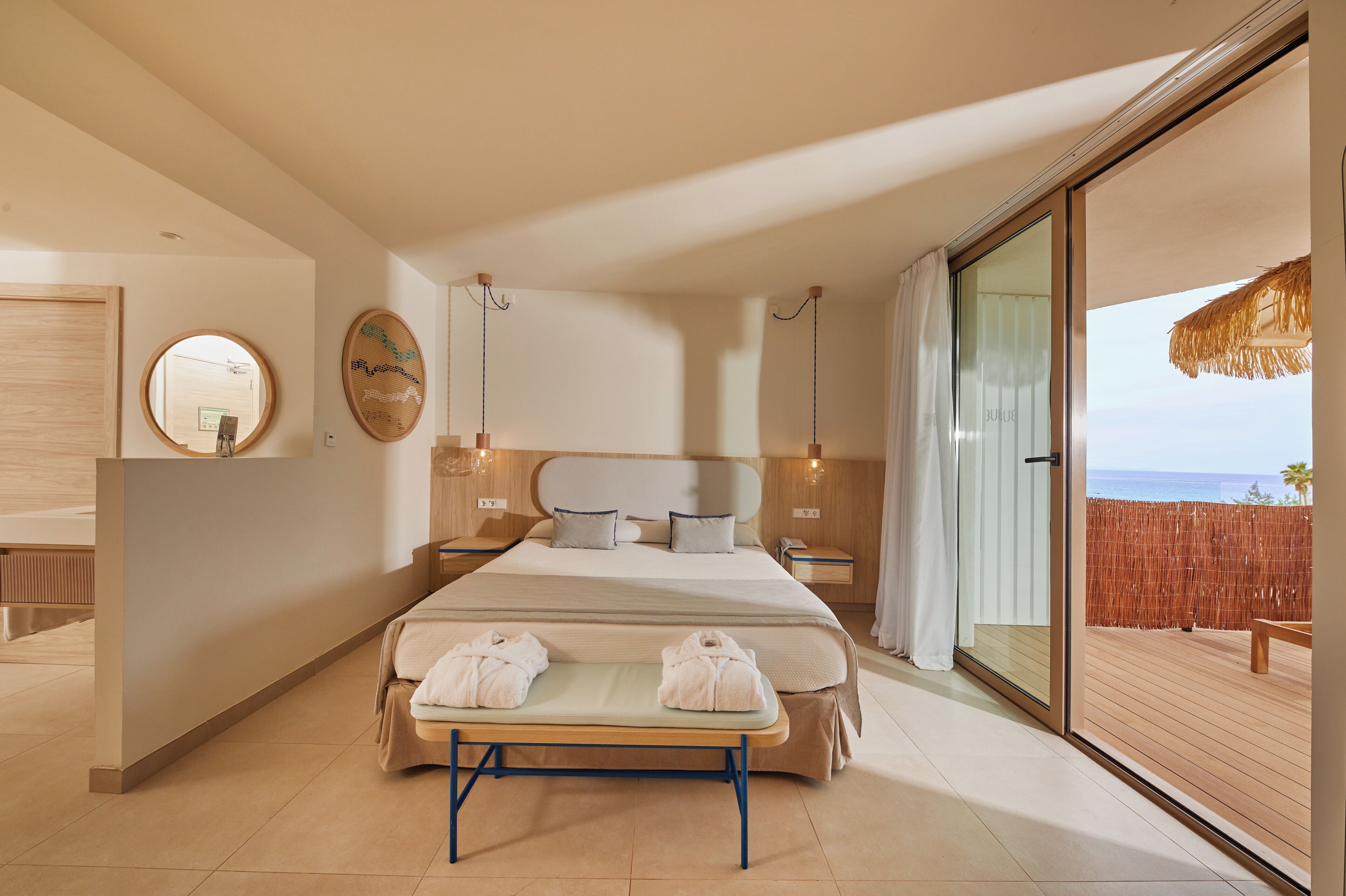 A modern hotel room with a large bed, a bedside table, a mirror, and a view of the sea through a sliding glass door.