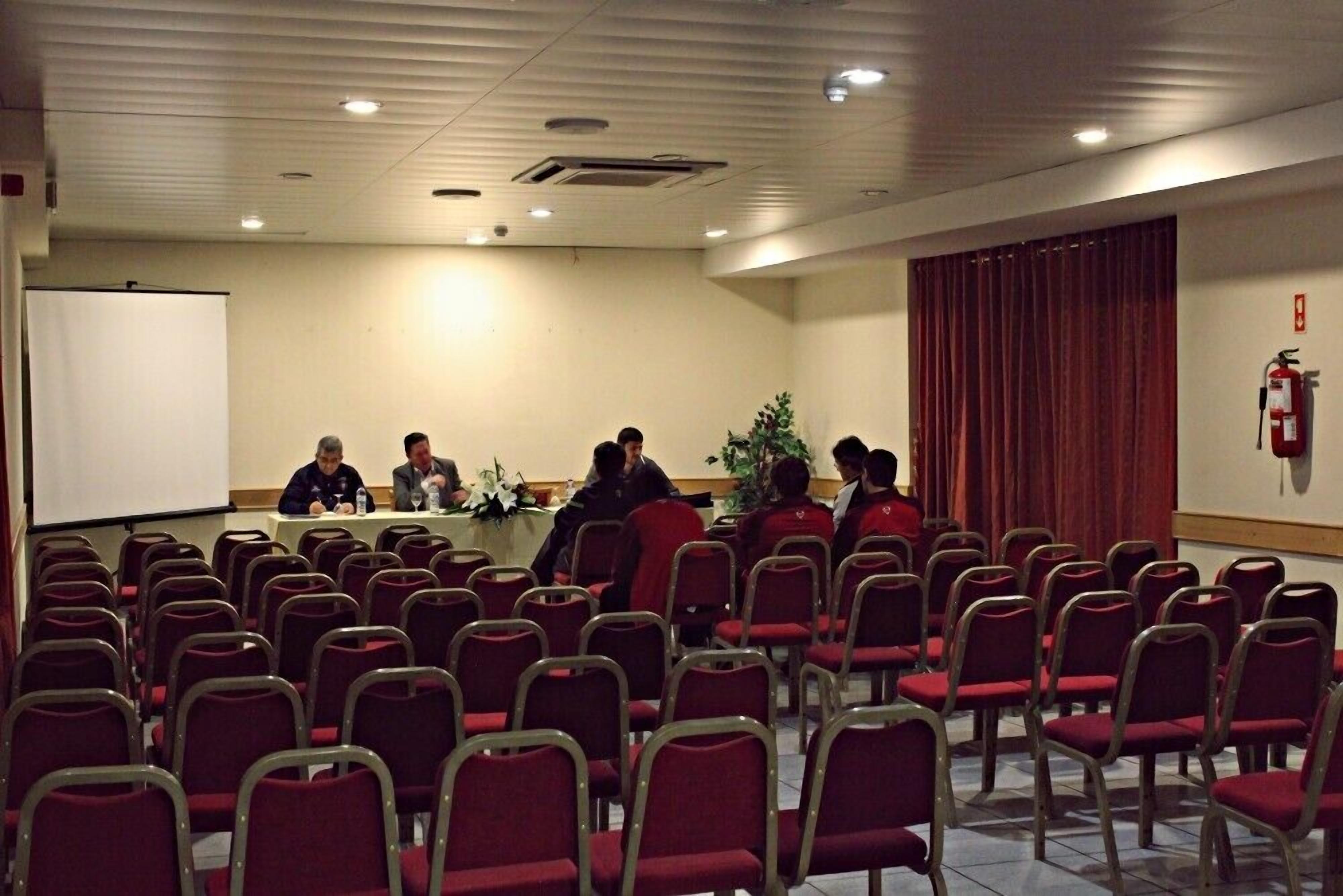 Ein Konferenzraum mit Reihen roter Stühle, einer Tafel, einer Projektionsleinwand und einem Feuerlöscher an der Wand.